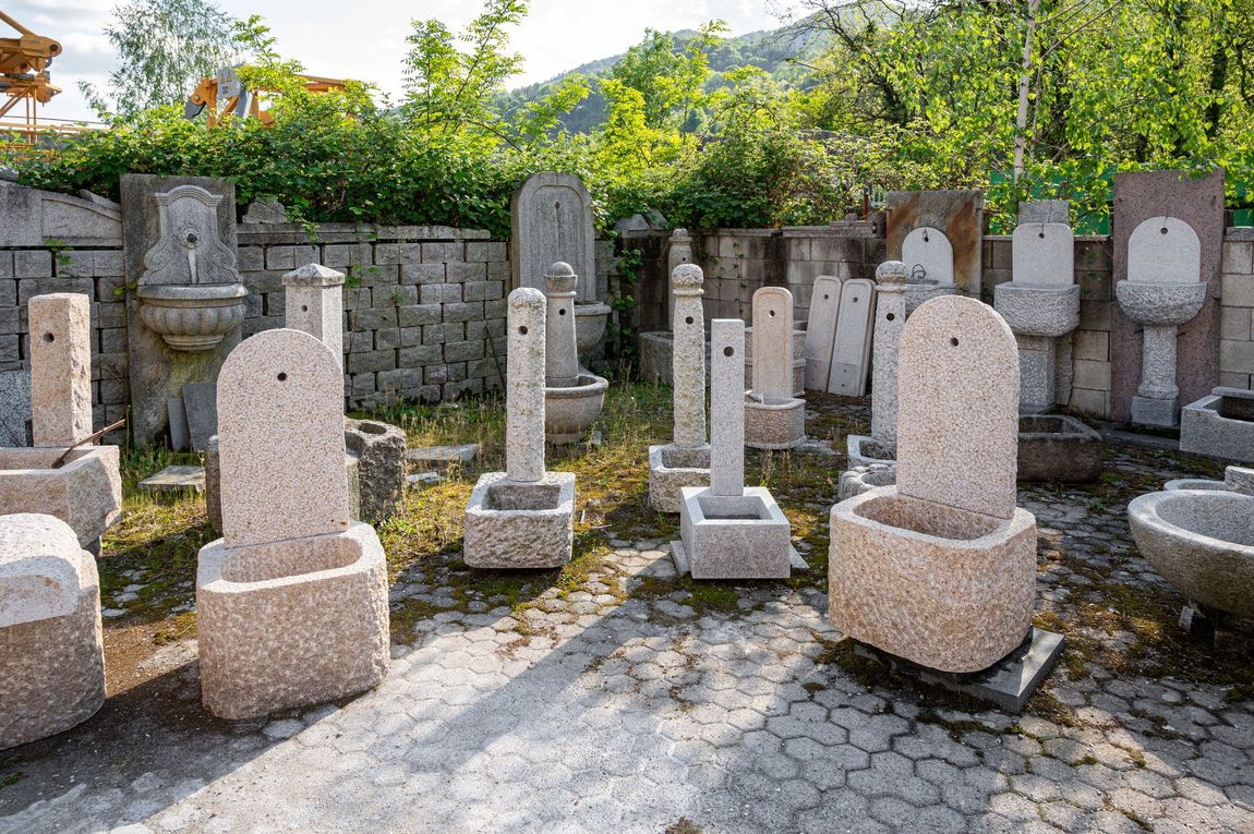 Una selezione di fontane e vasche in pietra scolpita, esposte in un cortile esterno in pietra con alberi sullo sfondo.