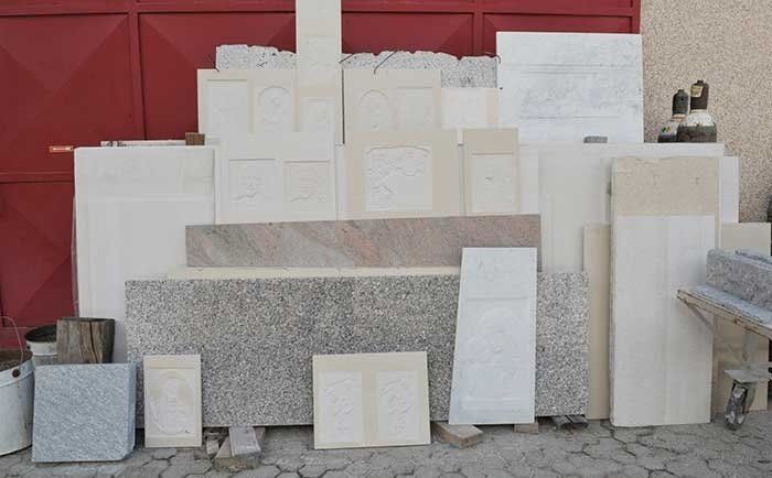 Una collezione di lastre e lapidi in granito e marmo di vario tipo, esposte contro una parete metallica rossa.
