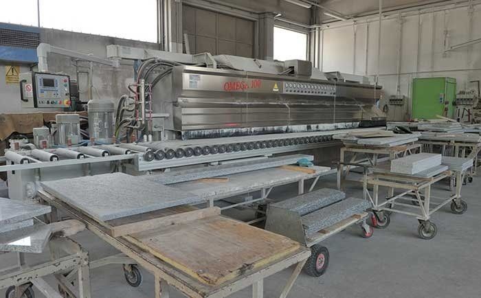 In un'officina è presente una macchina industriale per la lavorazione della pietra, dotata di rulli, con lastre di pietra su carrelli nelle vicinanze.