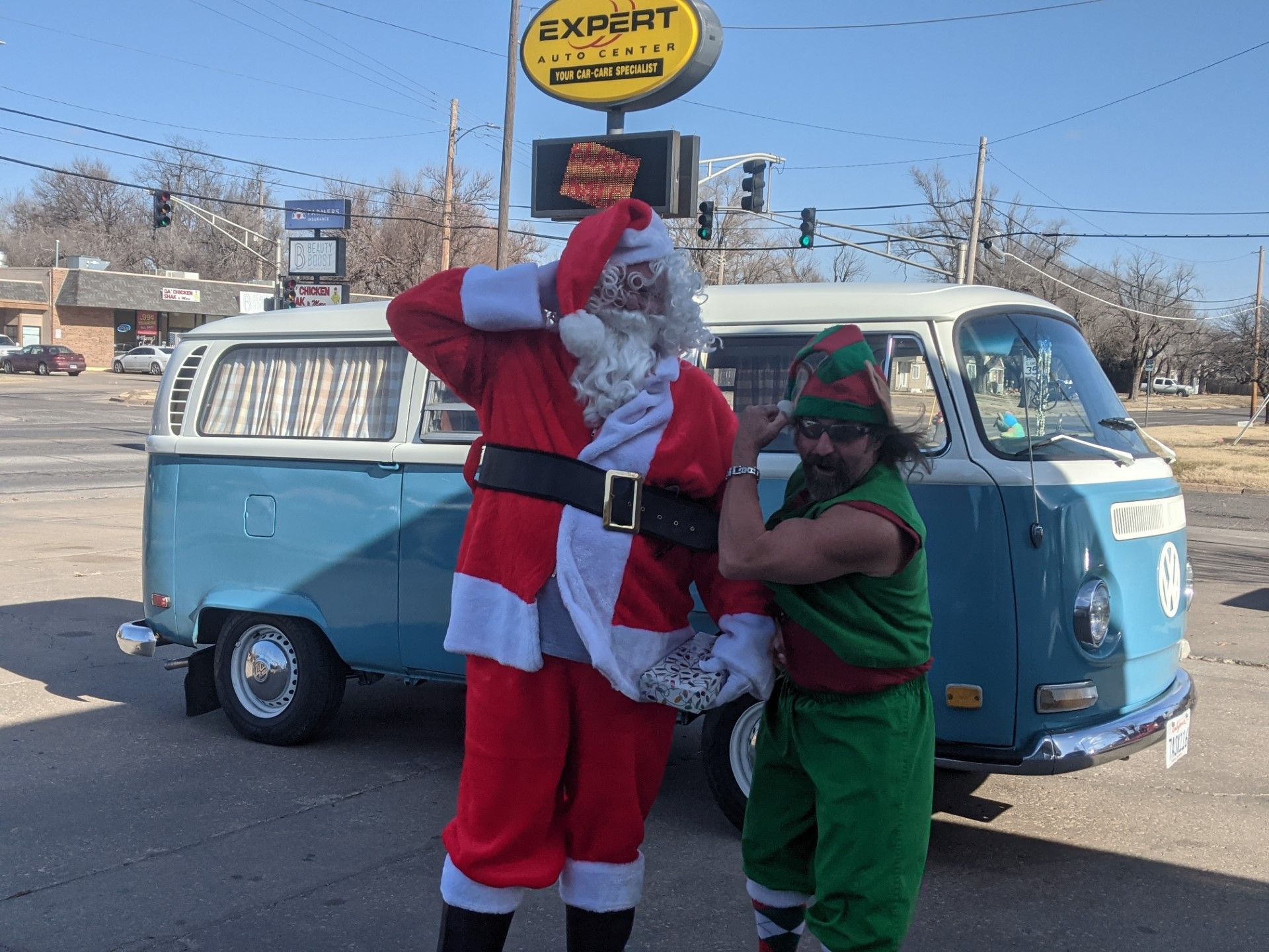 Santa and an elf posing in front of a blue van on a sunny street.