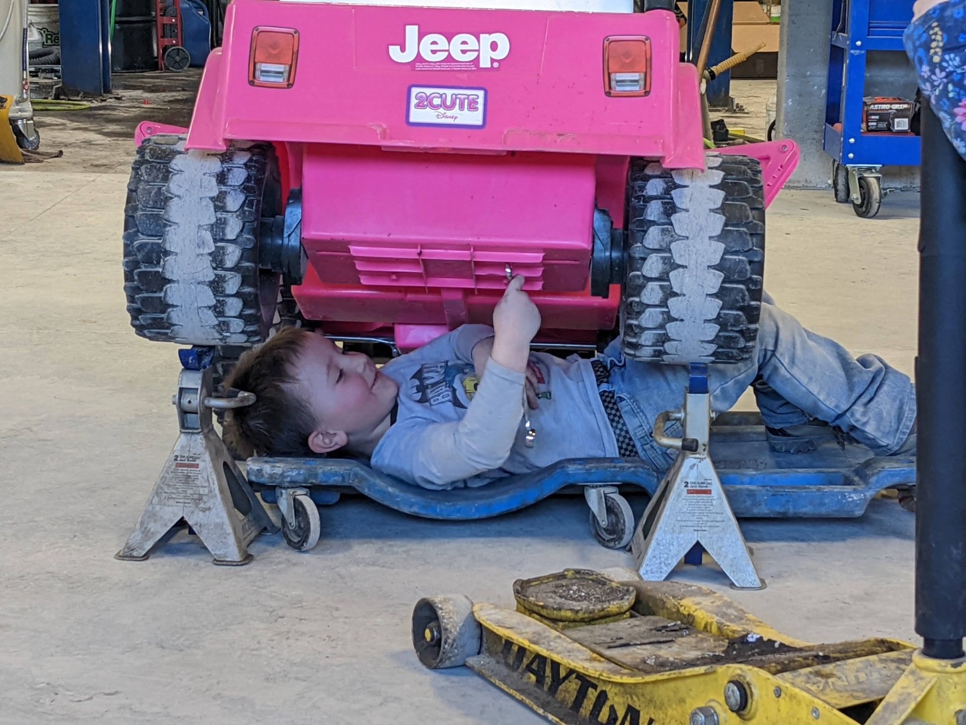 Mechanic lying under a pink Jeep, working beneath the chassis on a garage floor.