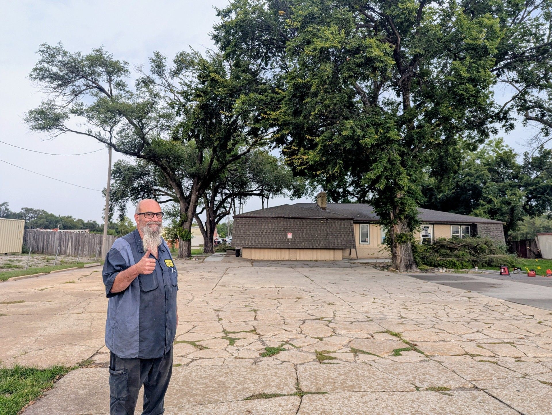 A man stands in a cracked stone courtyard near large trees and small buildings under a cloudy sky.