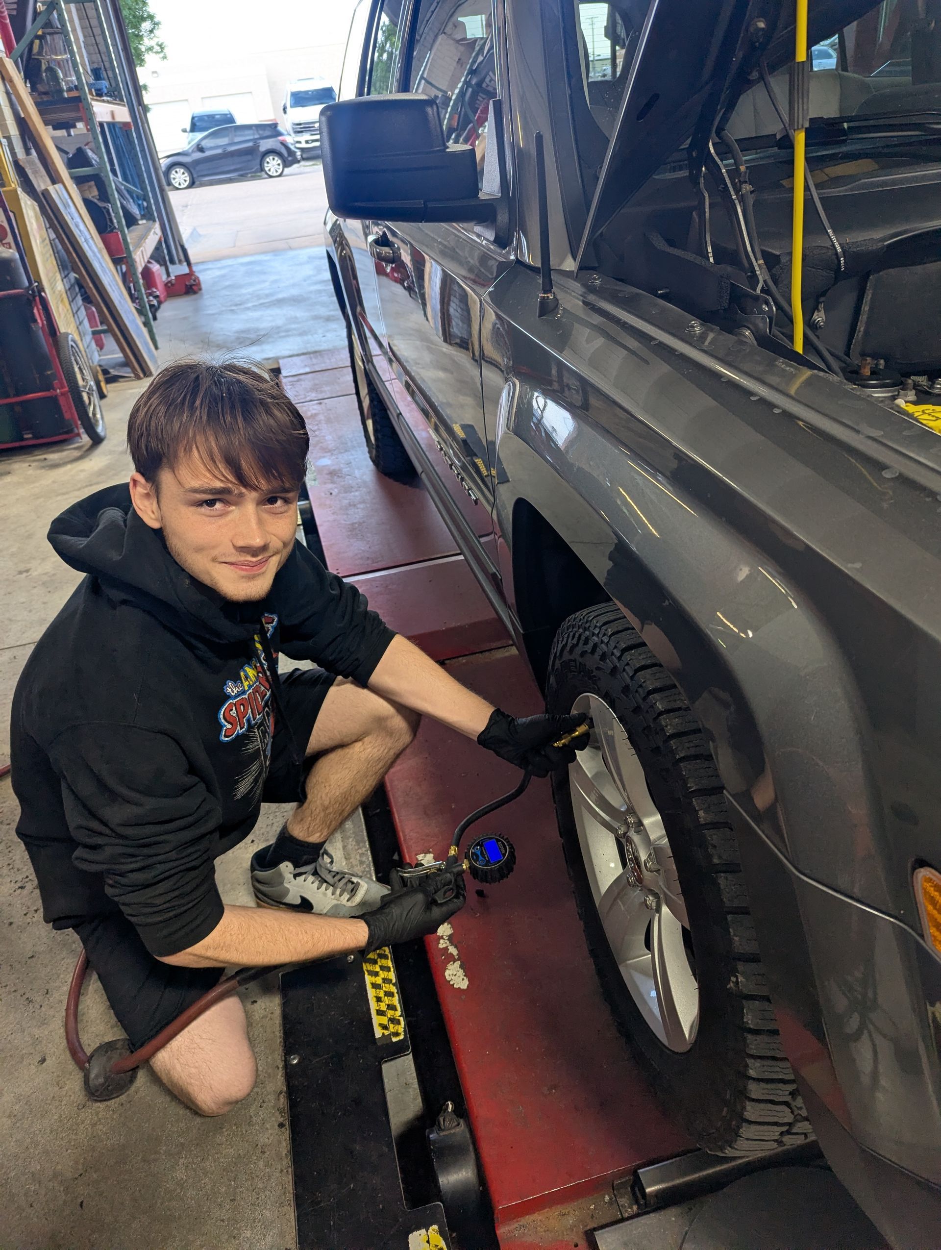 Mechanic kneels beside a gray SUV, using a tire tool in a garage bay.