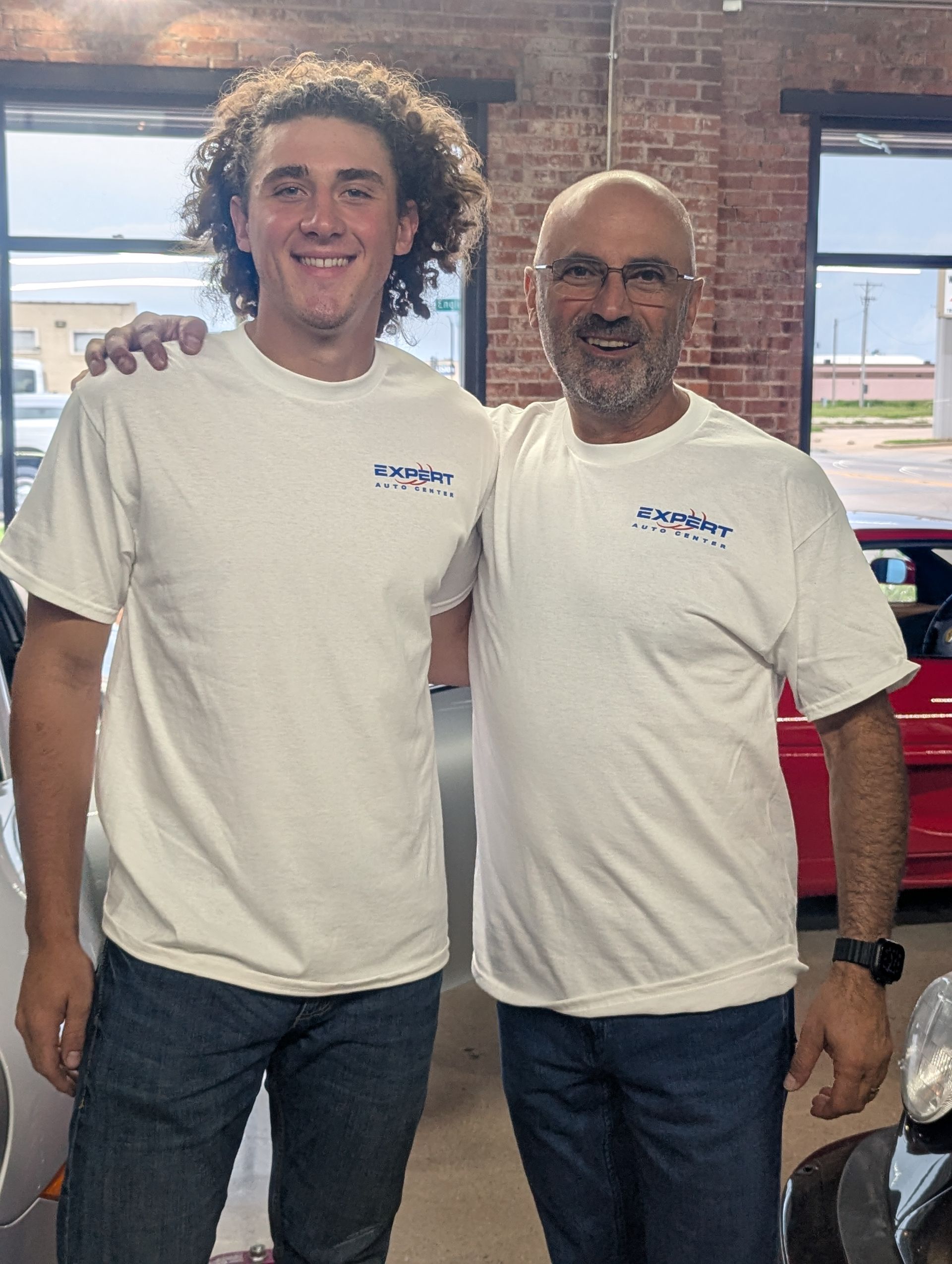 Two men in white T-shirts and jeans standing together indoors, smiling at the camera.