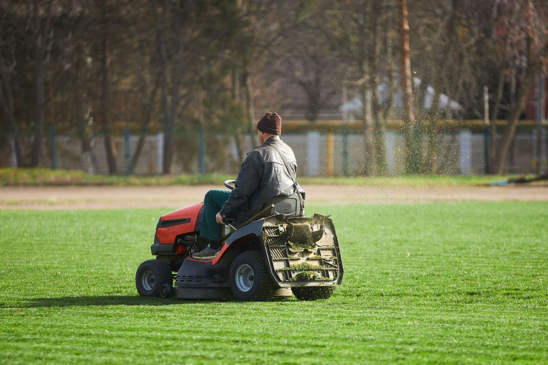 Person riding a red lawn mower on a green field, with trees in the background.