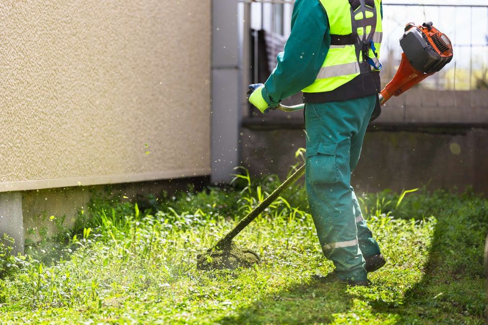 Person using a weed eater to trim grass near a beige building; wearing green work clothes and safety vest.