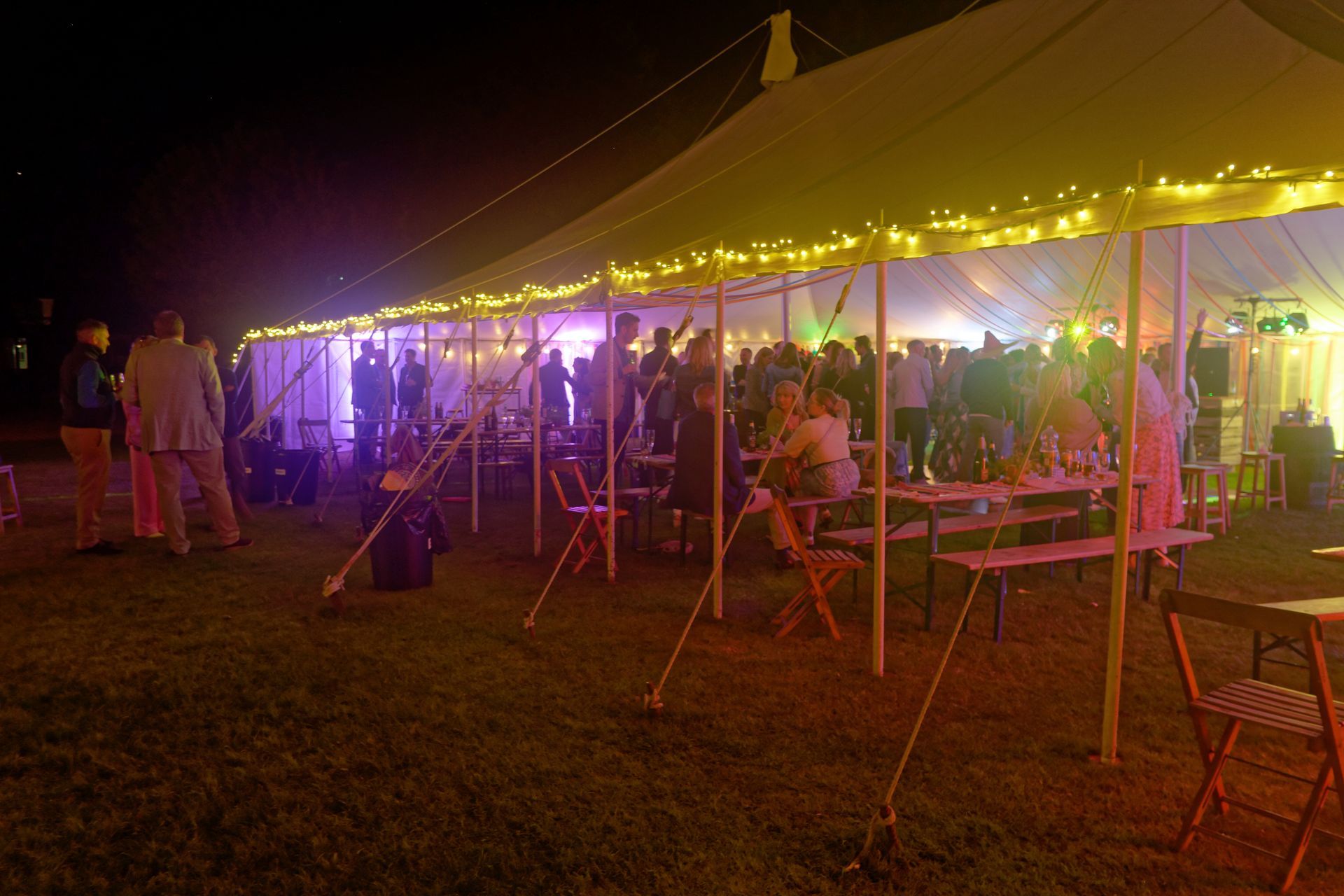 Party under a tent lit with string lights. People are mingling, sitting at tables, and enjoying the evening outdoors.