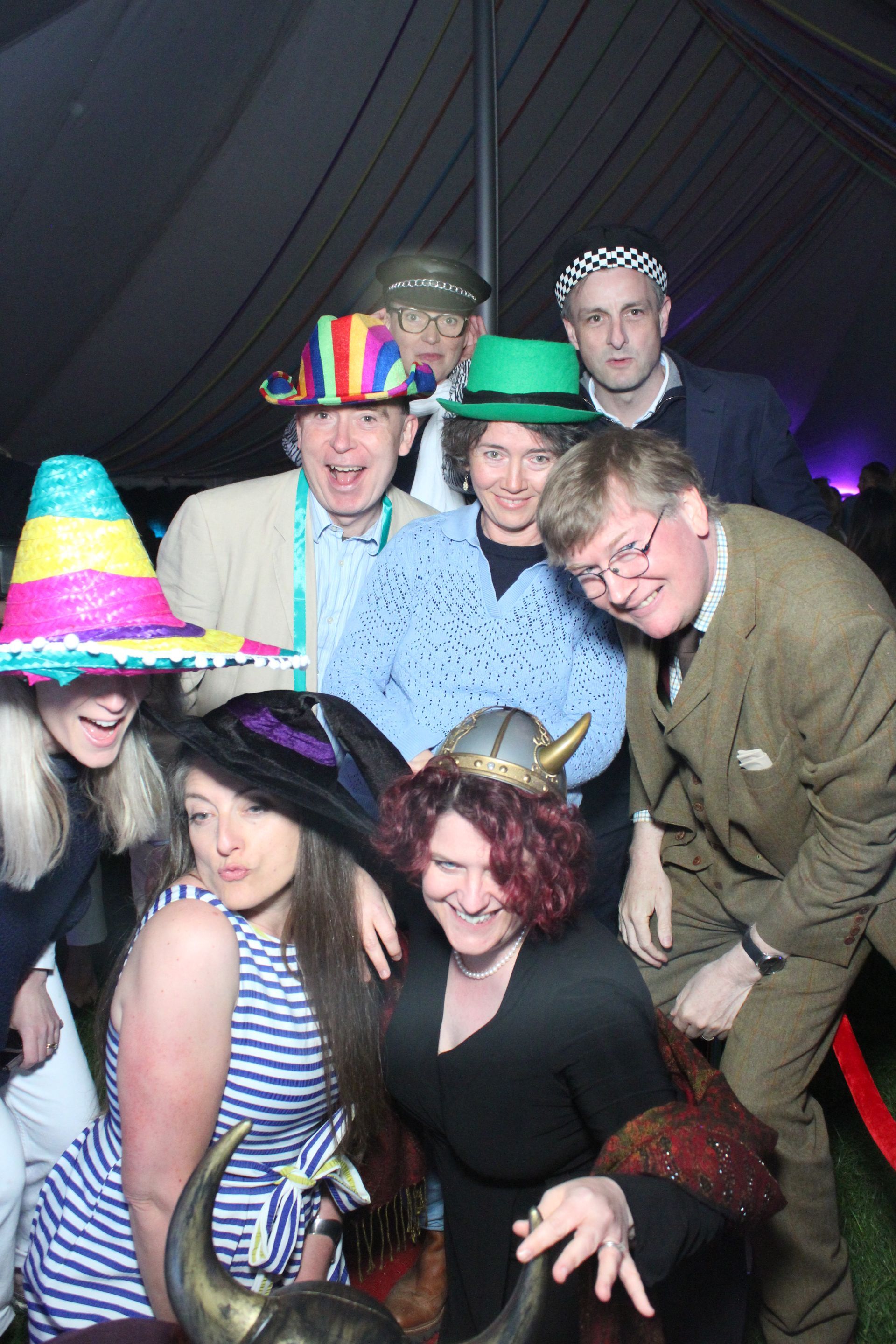 Group of people pose, wearing various hats, in a tent. Some are smiling, with a red carpet.