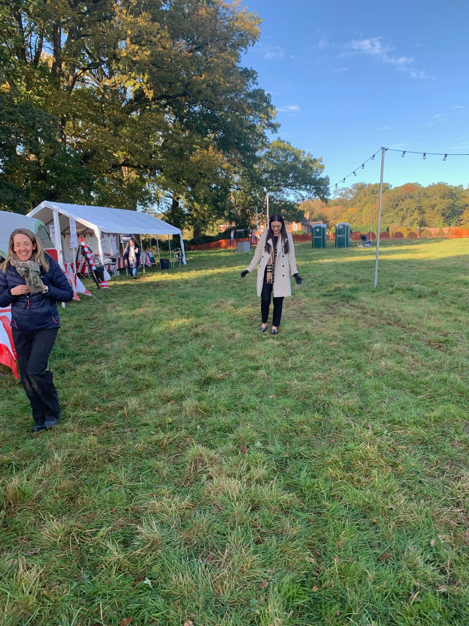 Two women walking in a grassy field, with tents and trees in the background on a sunny day.