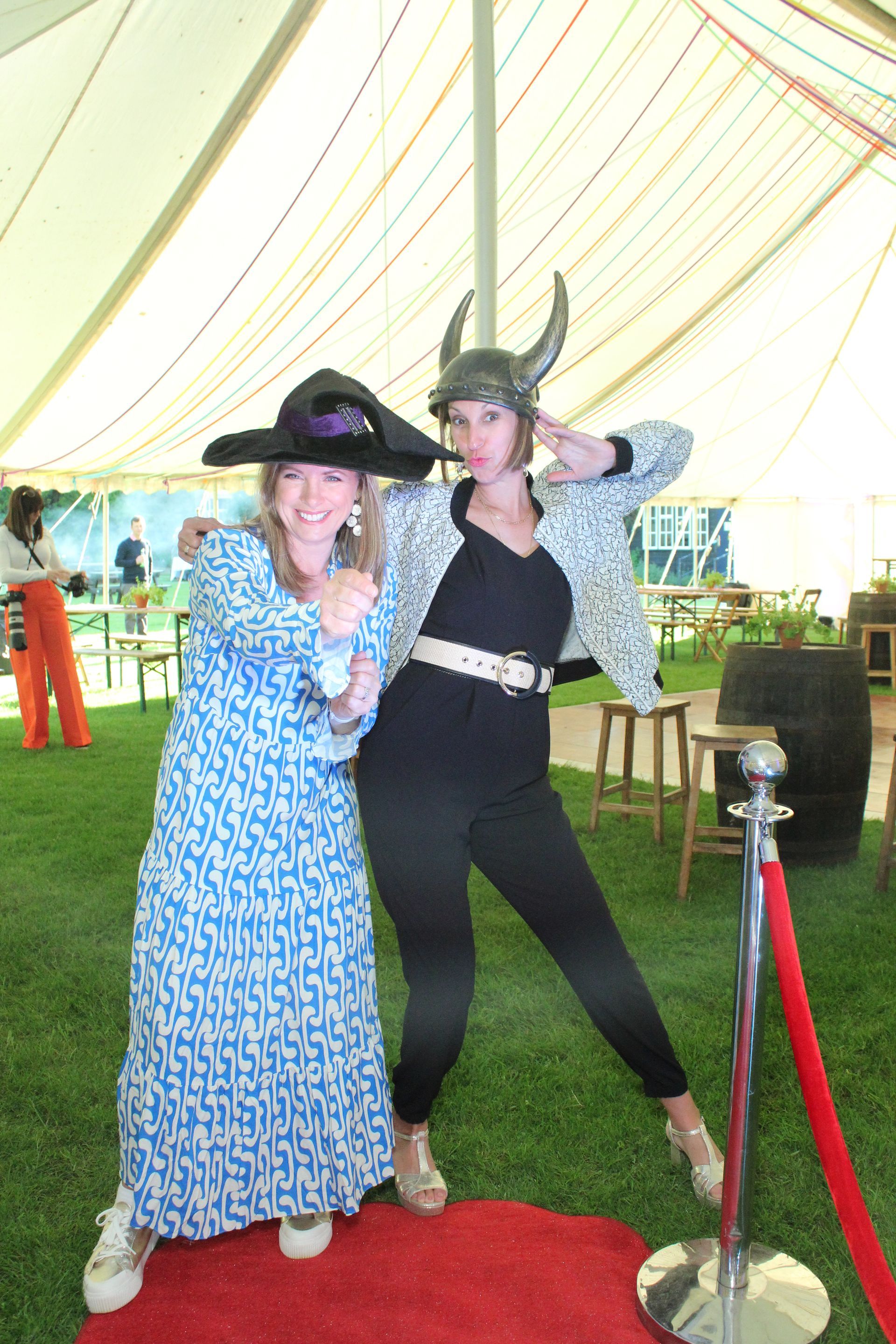 Two women in hats pose on a red carpet in a tent. One wears a Viking helmet, the other a wide-brimmed hat.