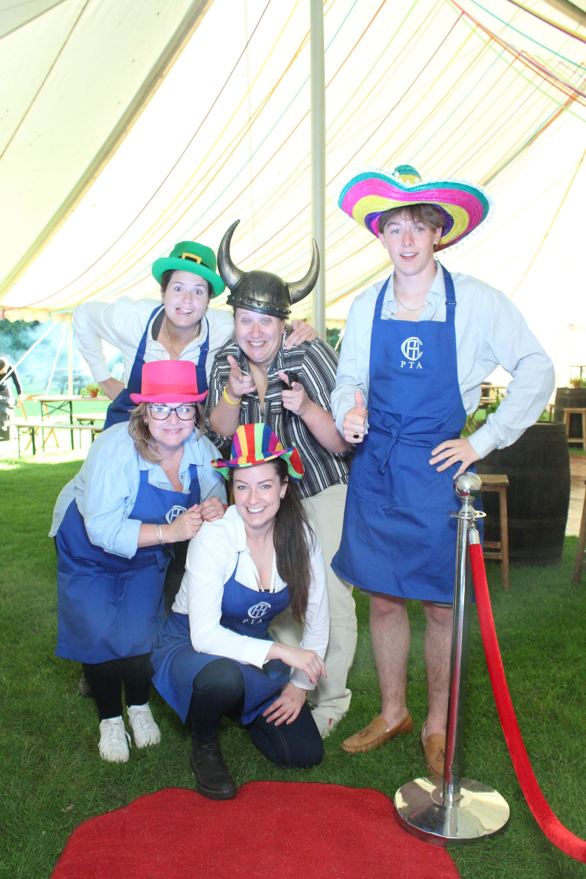Five people in hats and aprons, posed on a red carpet, under a tent on grass.