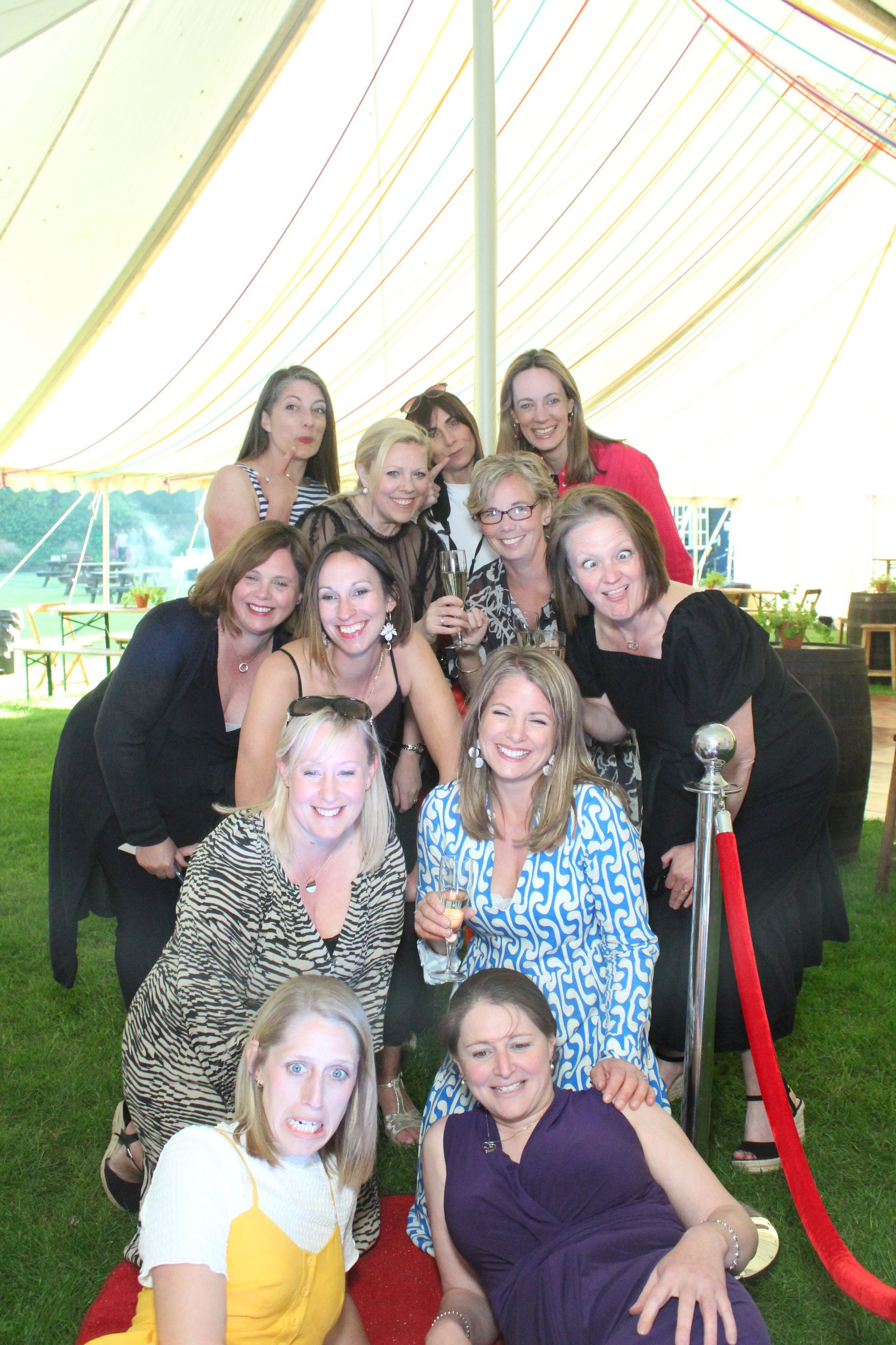 Group of women posing together, some with drinks, on red carpet, outdoors.