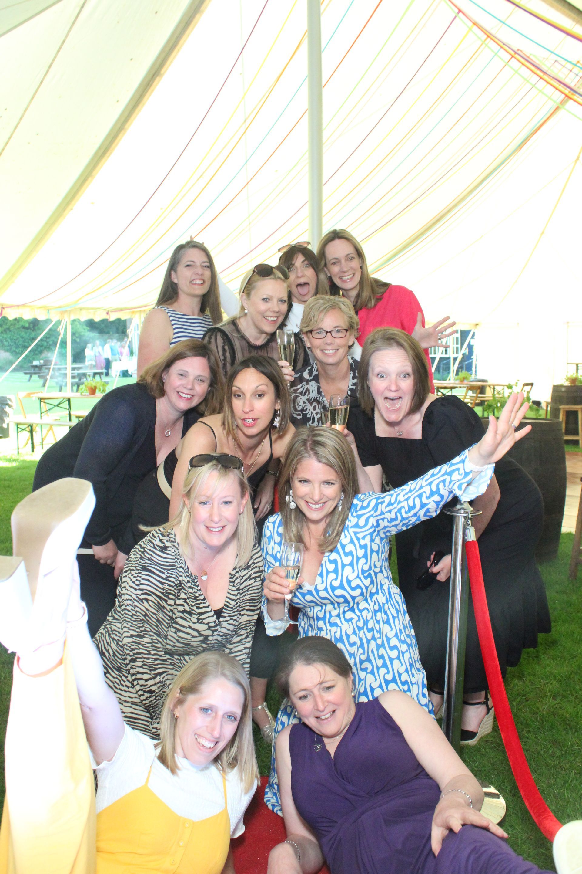 Group of women smiling, posing on a red carpet under a tent.