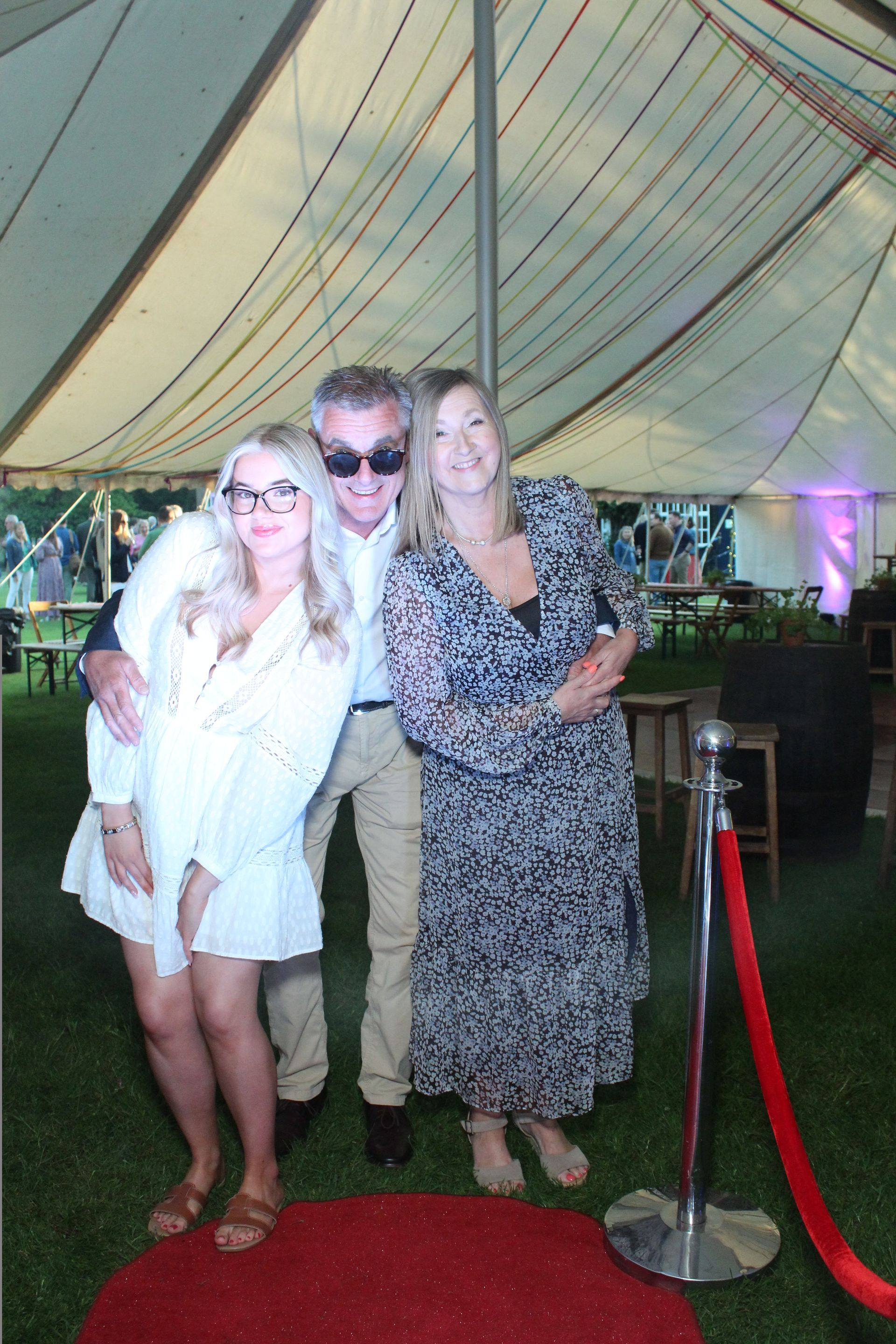 Three people pose by a red carpet inside a tent. Woman in white hugs man, woman in floral dress smiles.