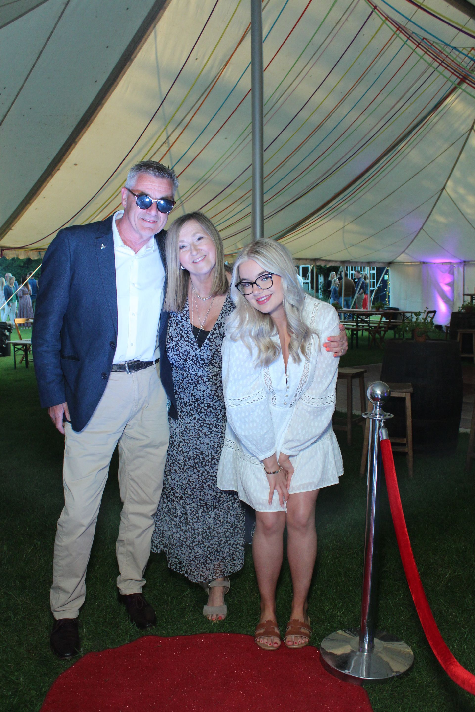 Three people pose on a red carpet under a tent. A man in a blazer, woman in a dress, and woman in a white outfit smile.