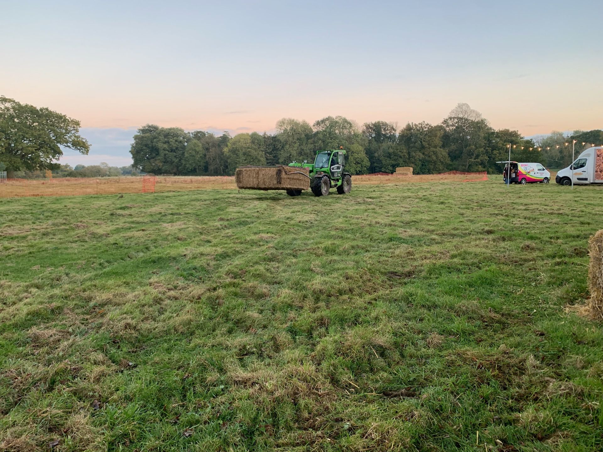 Green tractor with a hay trailer in a field, gathering hay bales at dusk.