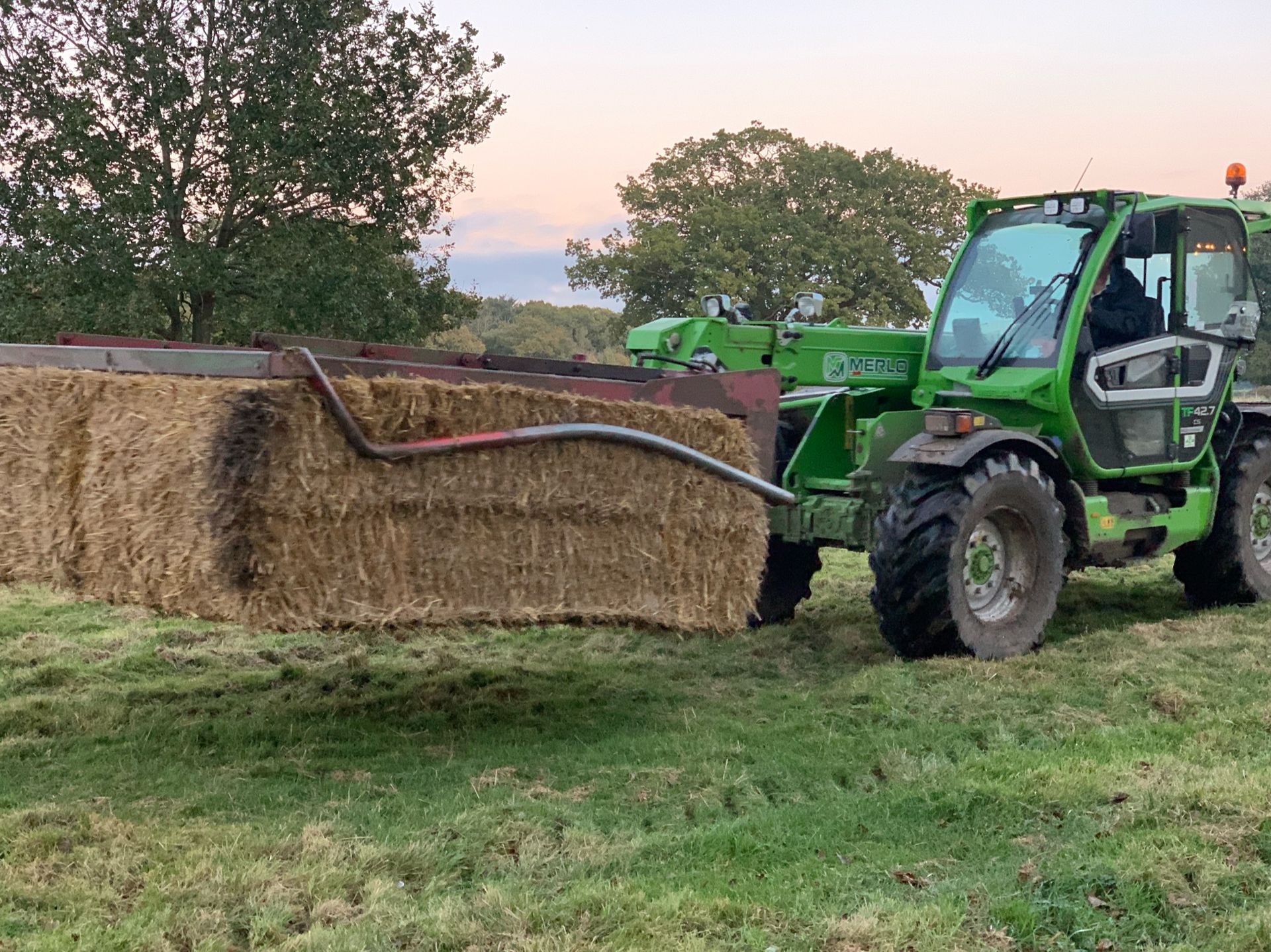 Green telehandler carrying a large rectangular bale of hay in a grassy field.