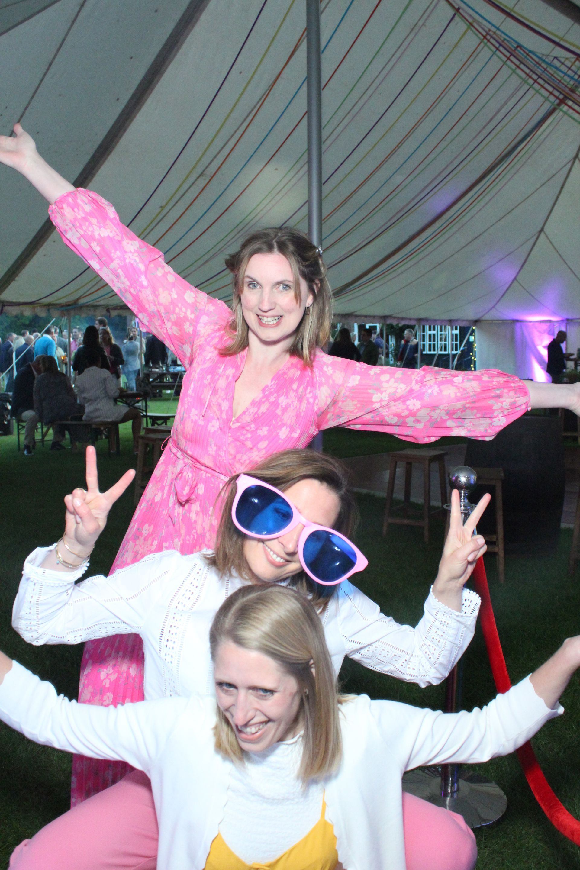 Three women posing in a photo booth, arms raised in fun. One in pink, others in white, all smiling.