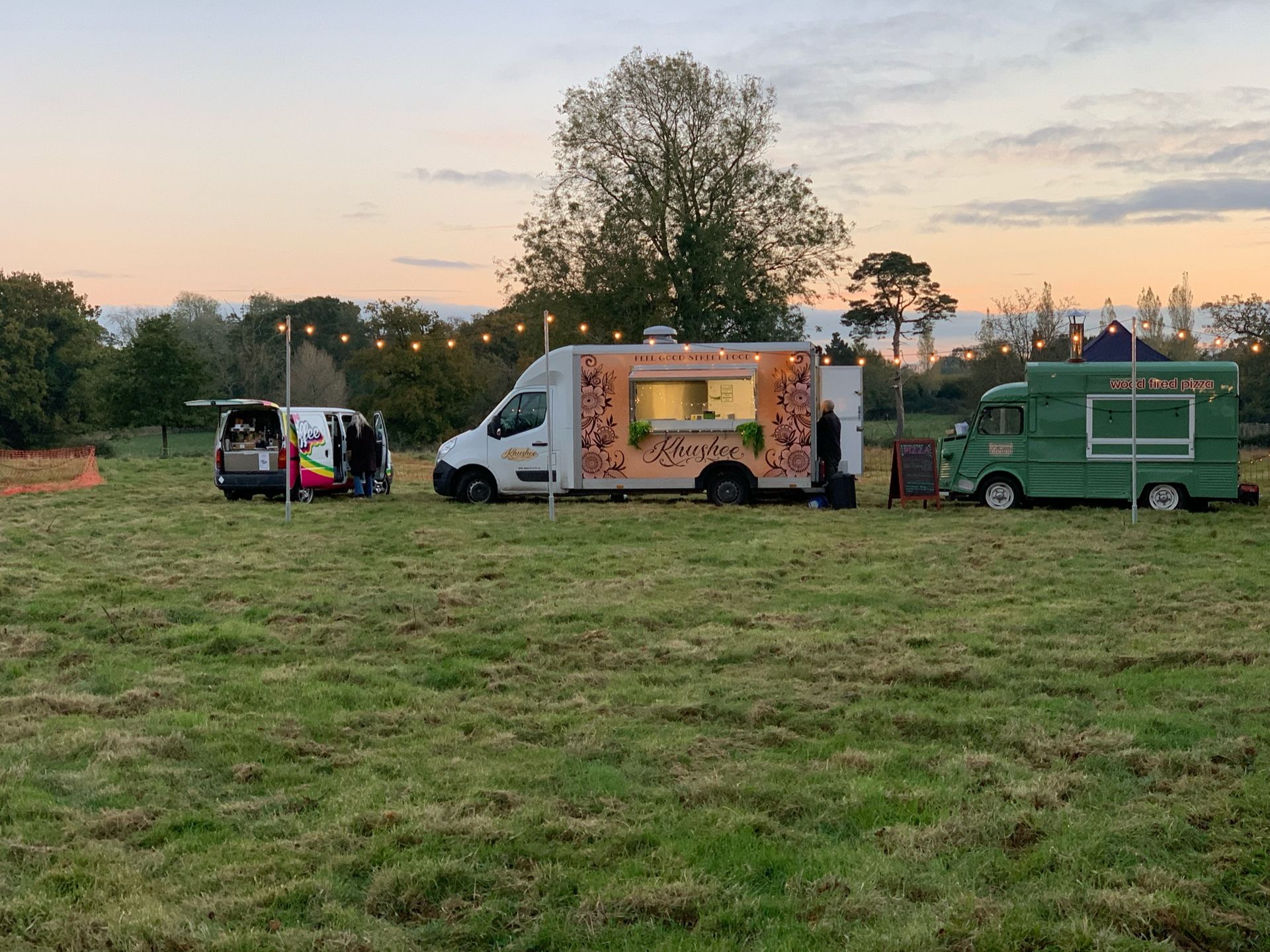 Three food trucks parked on a grassy field. One white, one green, one black. Strings of lights overhead. Sunset.