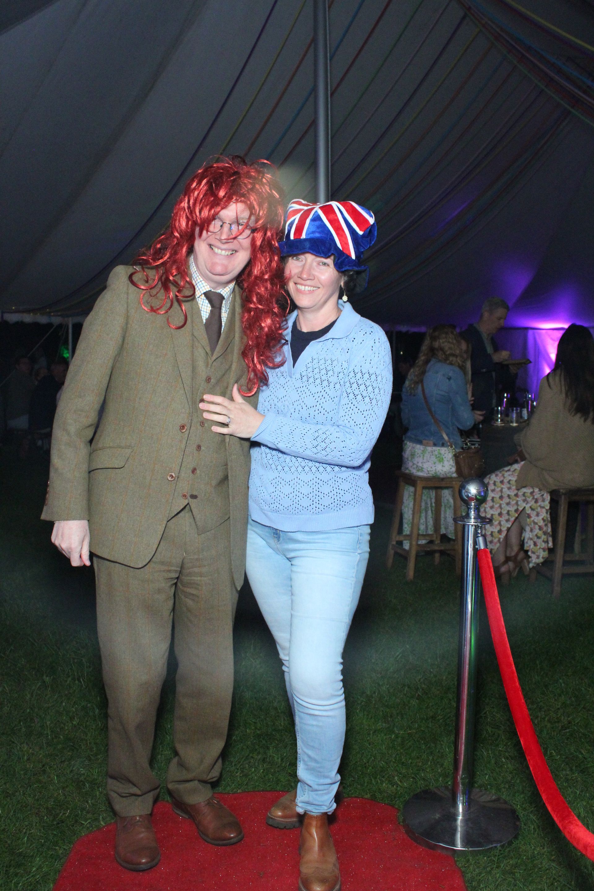 Two people posing on a red carpet. Man in a suit and wig, woman in a Union Jack hat and jeans.