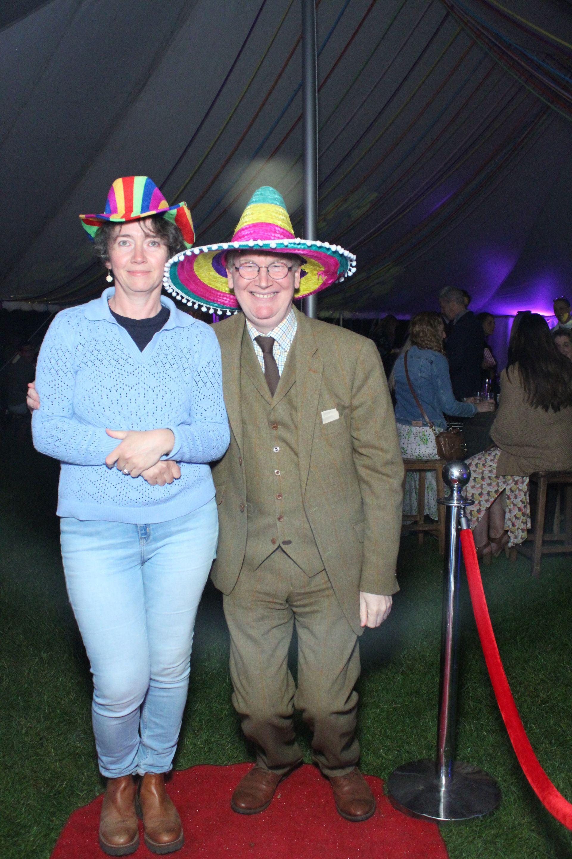 Two people in rainbow sombreros pose on a red carpet.