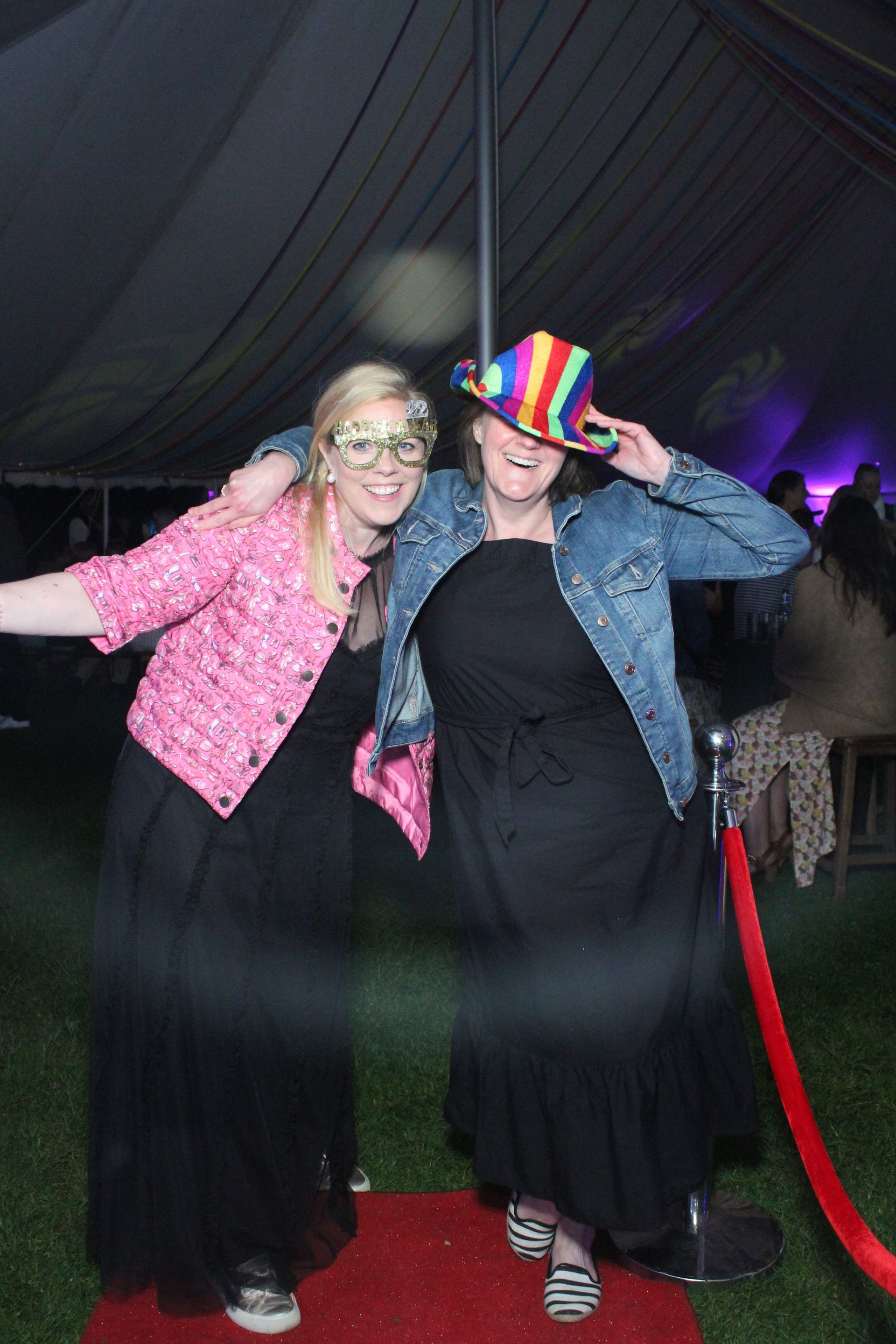 Two women posing on a red carpet, one in a pink jacket and the other in denim with a rainbow hat.