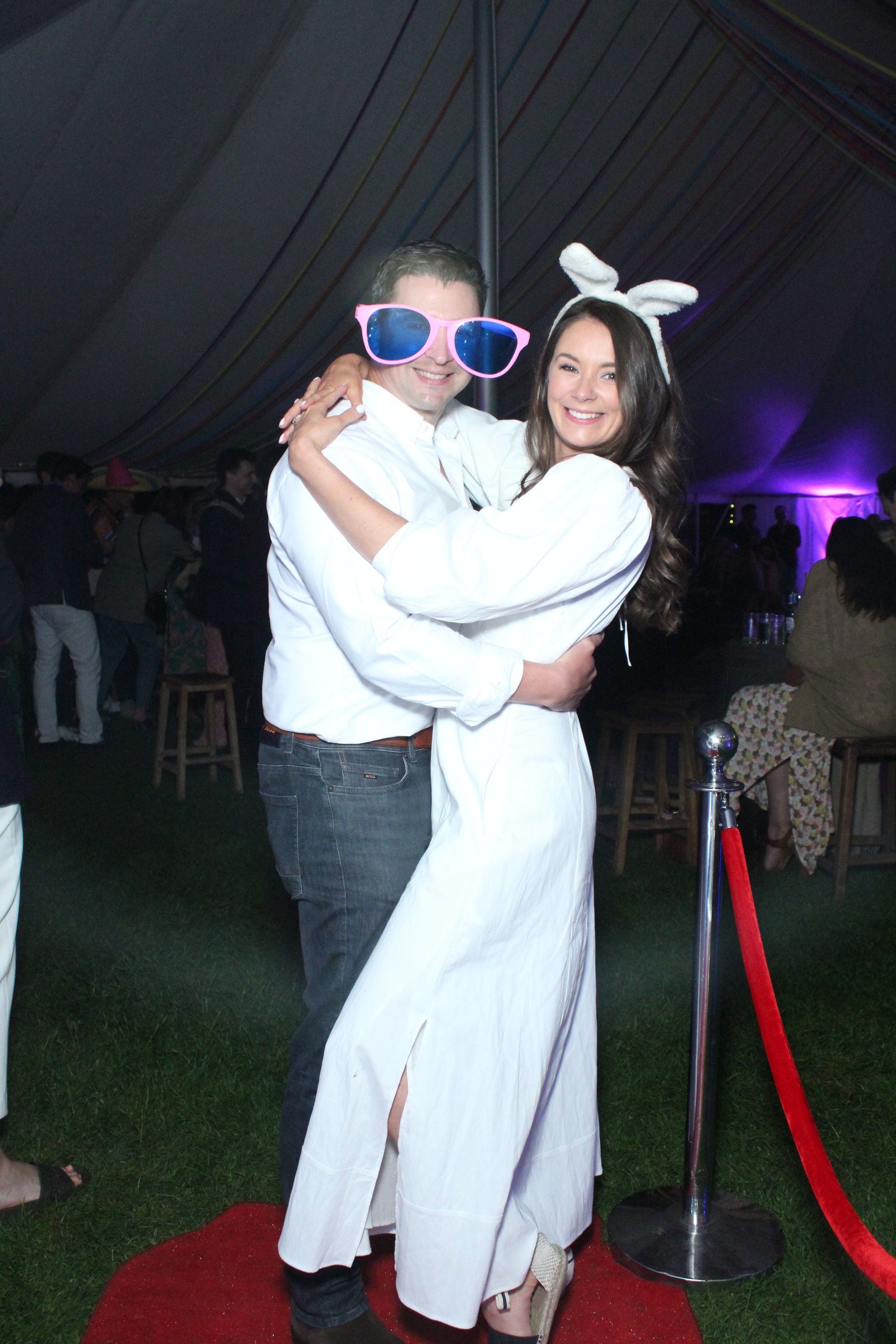 Couple dancing, man in big sunglasses, woman with bunny ears, on red carpet. Tent background.