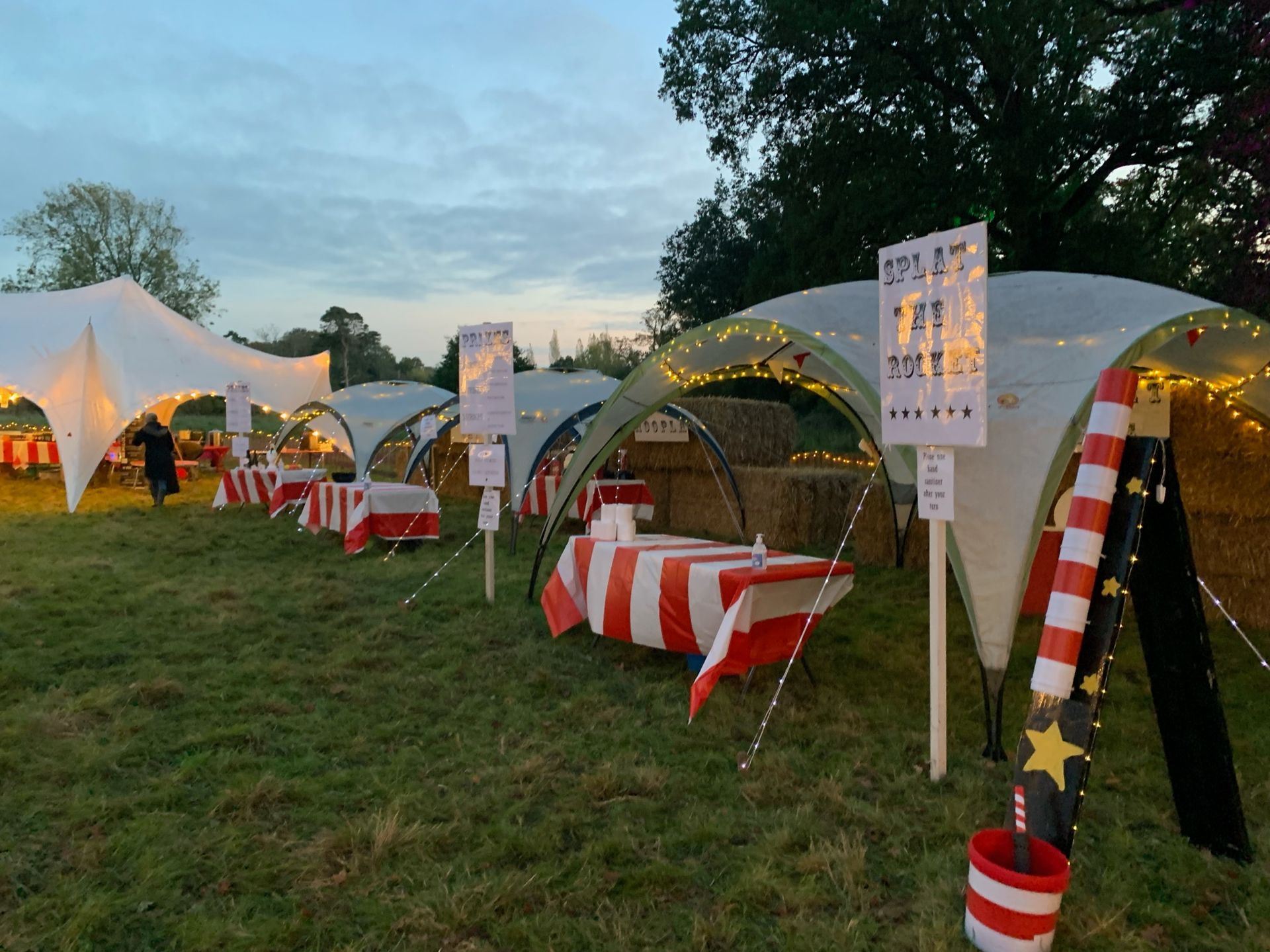 Tented booths with red and white striped tables at an outdoor event in a grassy field at dusk.