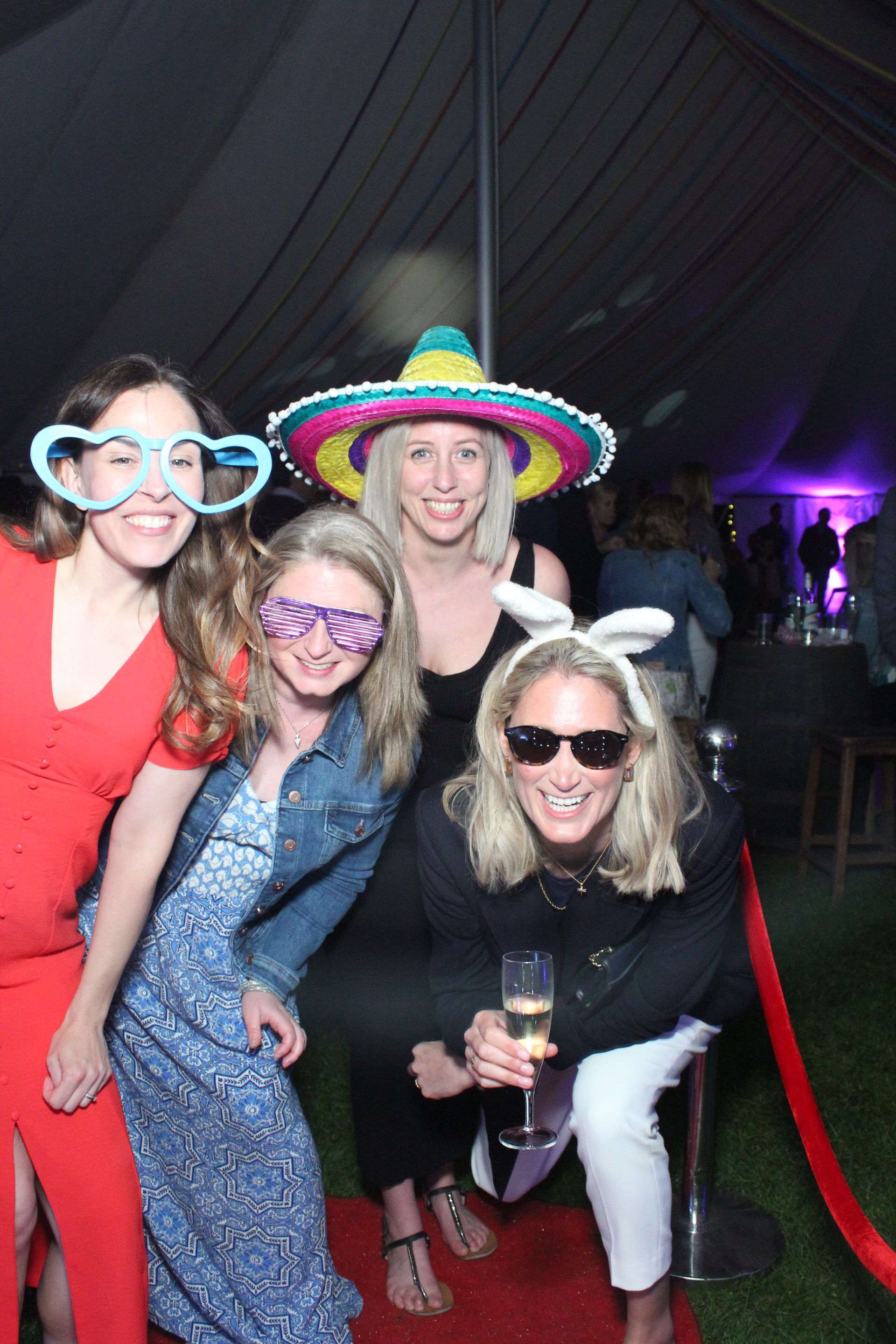 Four women pose for a photo. They wear costume glasses and hats at a red carpet event.