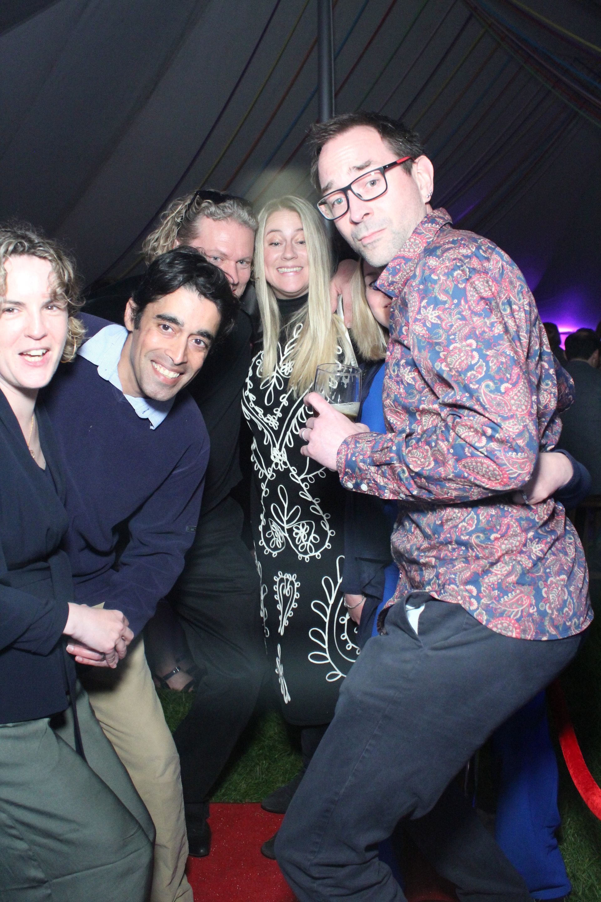 Group of friends posing for a photo, smiling and hugging in a tent with a red carpet.