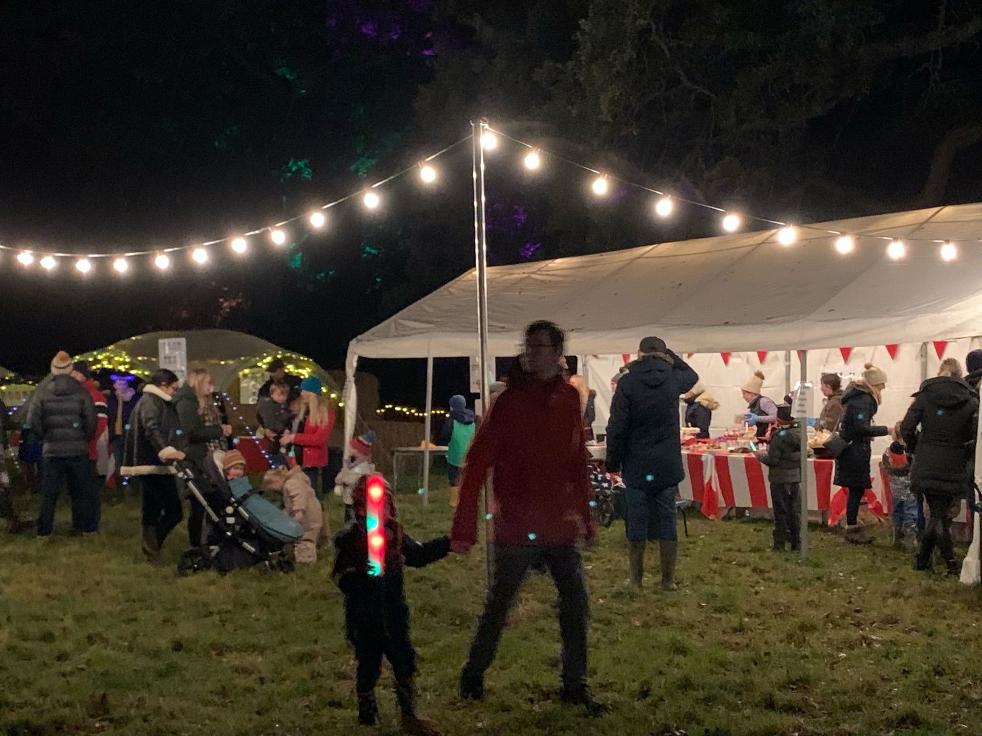 People at a night market with string lights; a man and child walk past a stall.