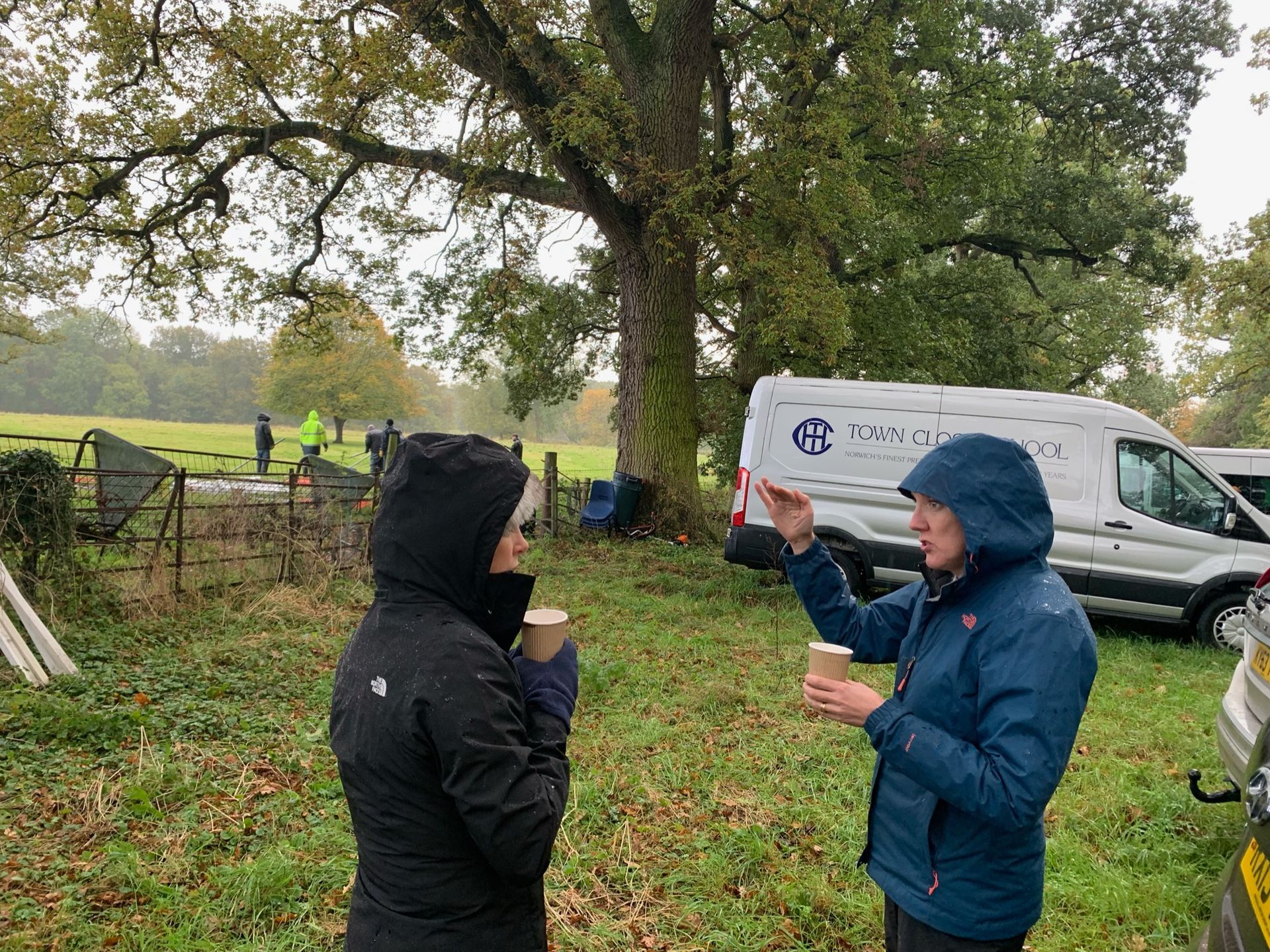 Two people in coats outdoors talking, one gesturing. A van and tree are in the background.