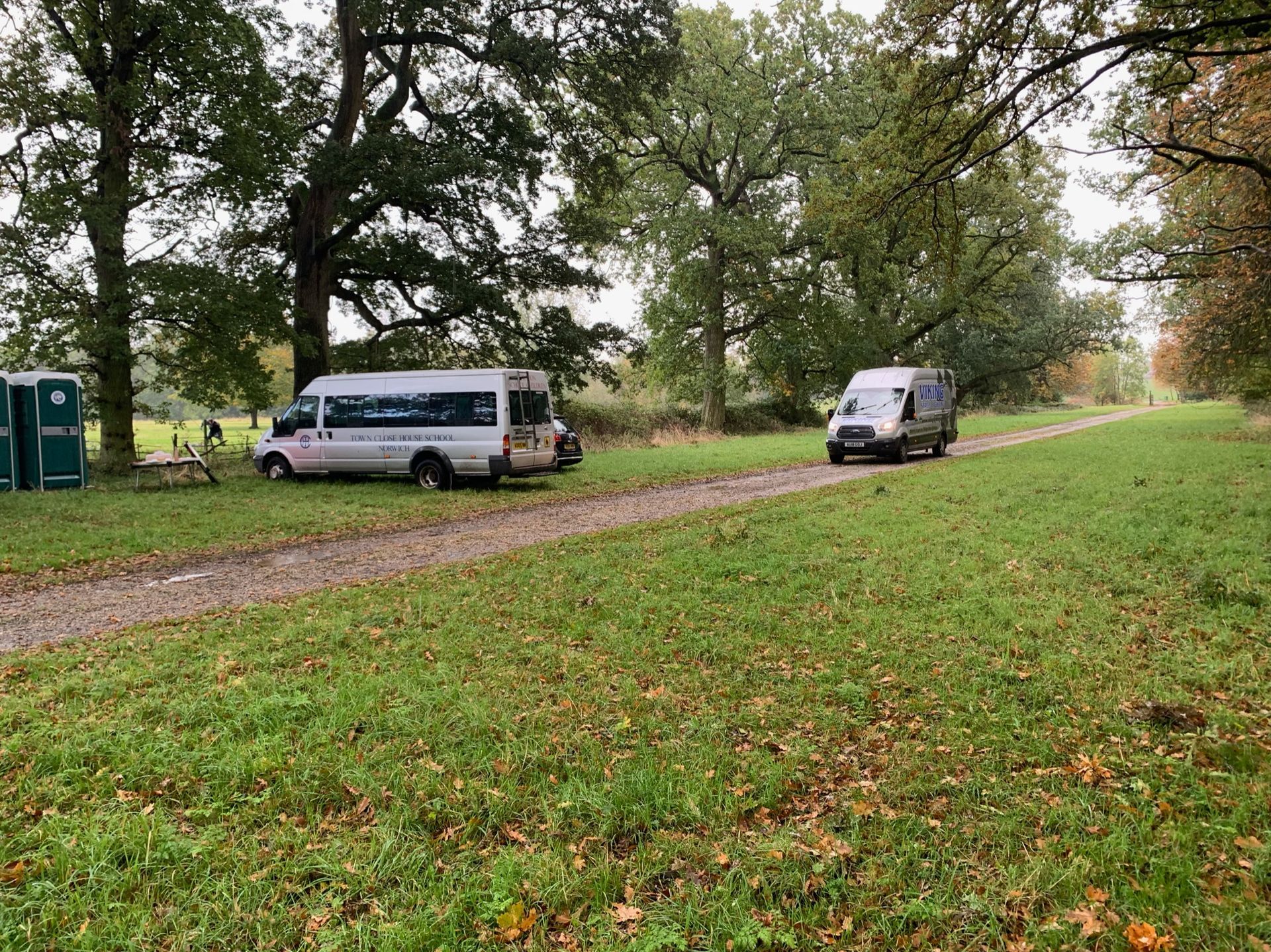 Two vans on a grassy path in a wooded area, with portable toilets visible to the left.