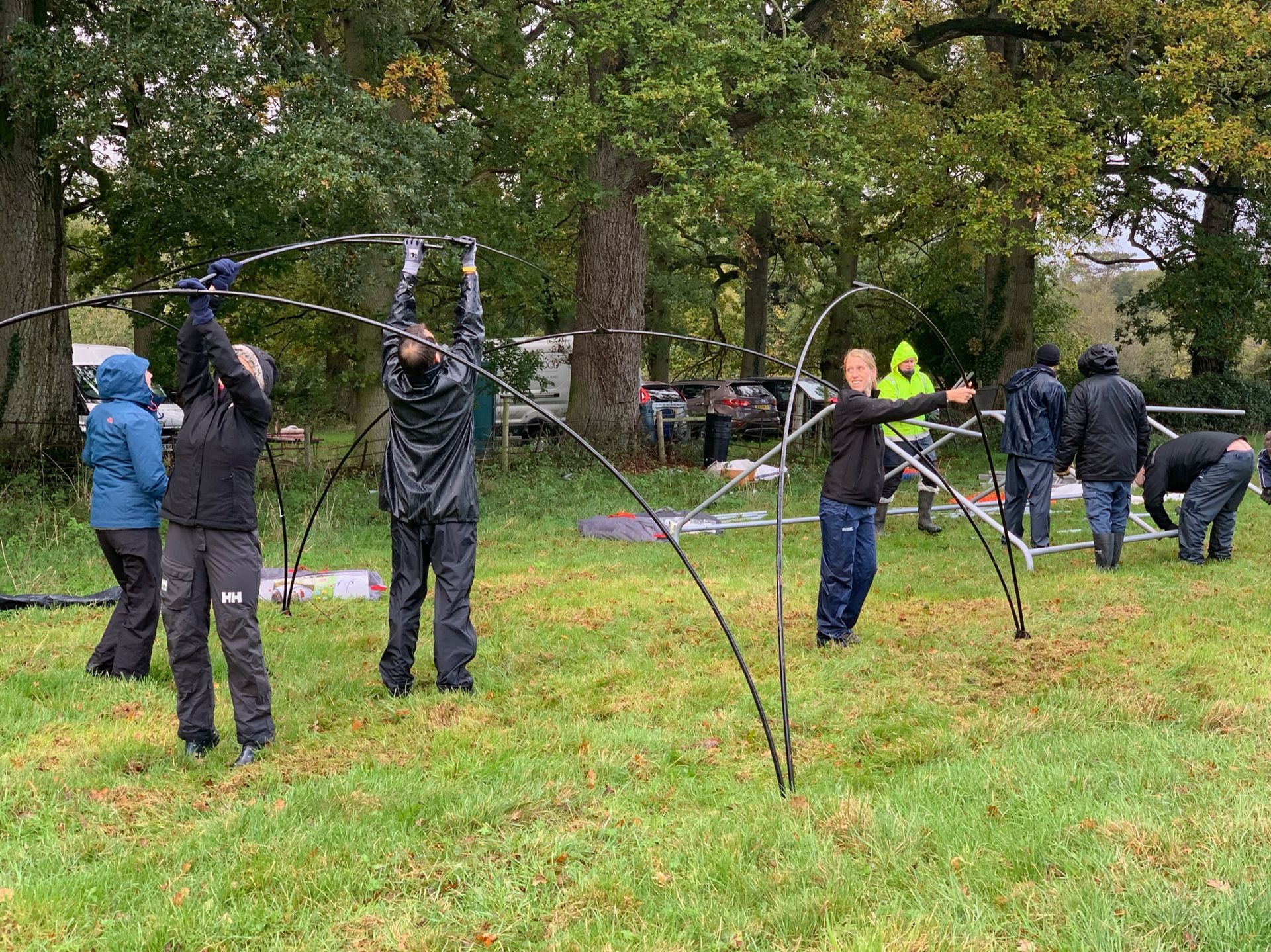 People assembling a curved structure outdoors on a grassy field. Overcast sky; trees in the background.
