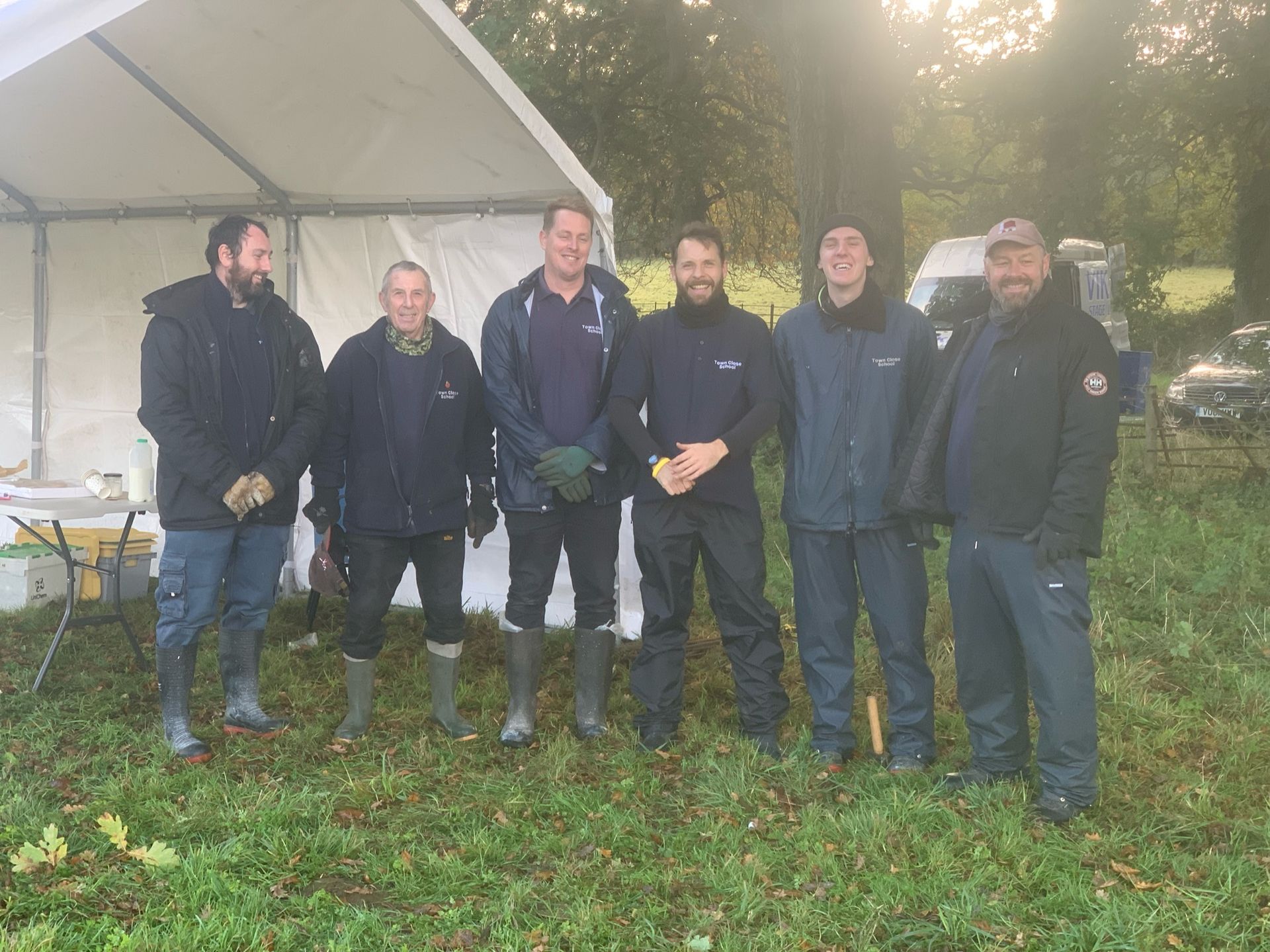 Six men standing in a grassy field under a tent. They wear outdoor clothing, and the sun shines in the background.