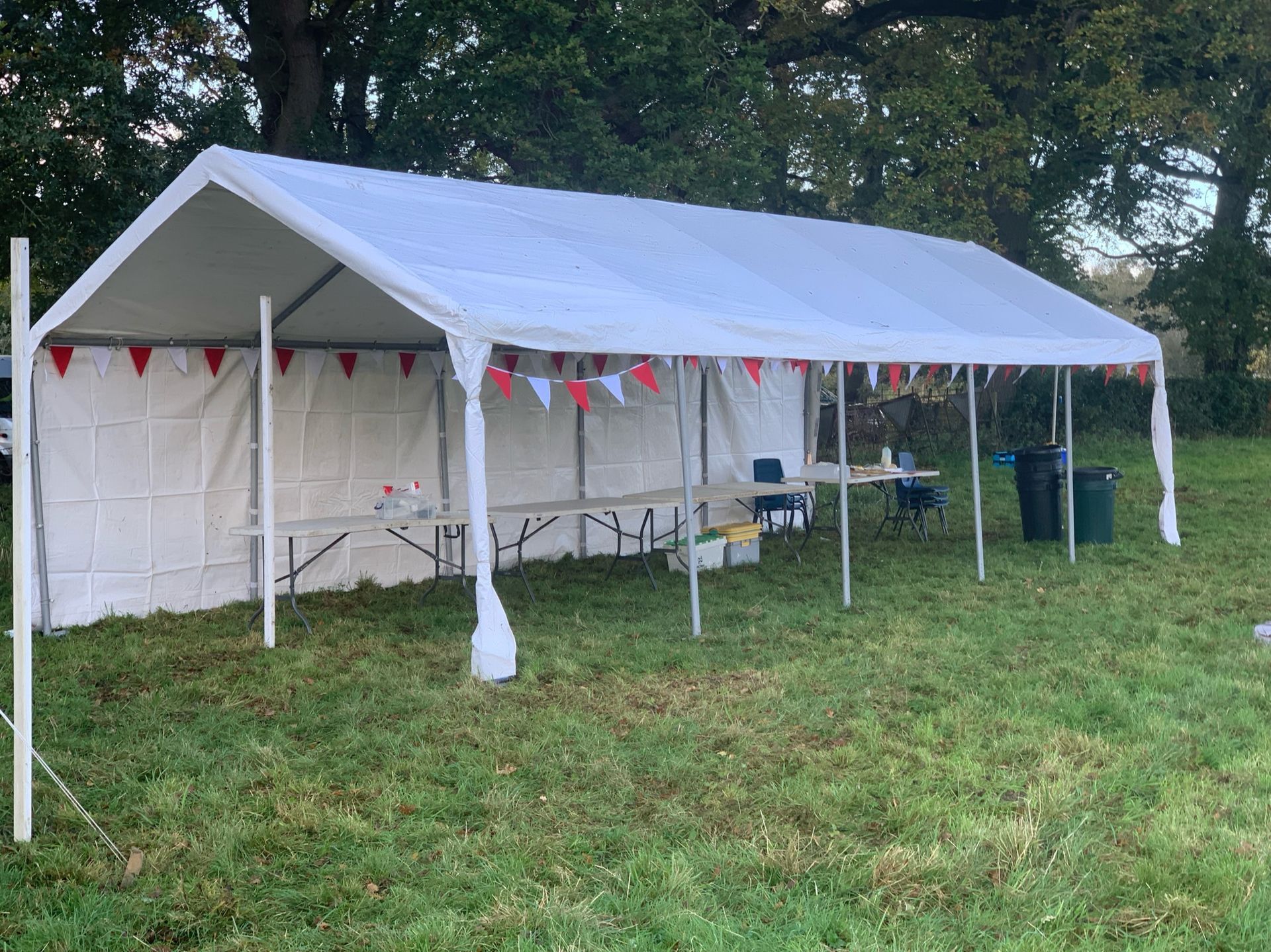 White tent on a grassy field; red and white bunting hangs from the roof; table and trash bin inside.