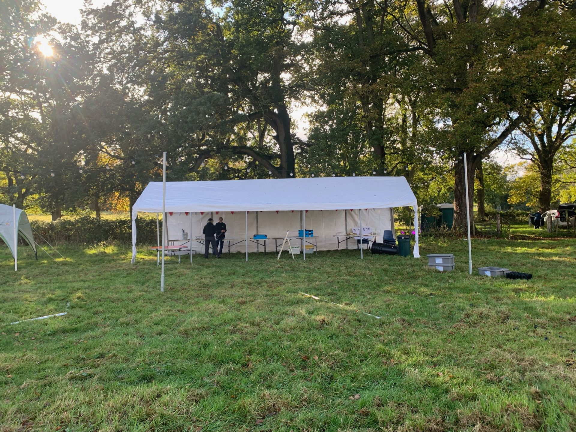 White tent in a grassy field with people standing inside, trees in the background.