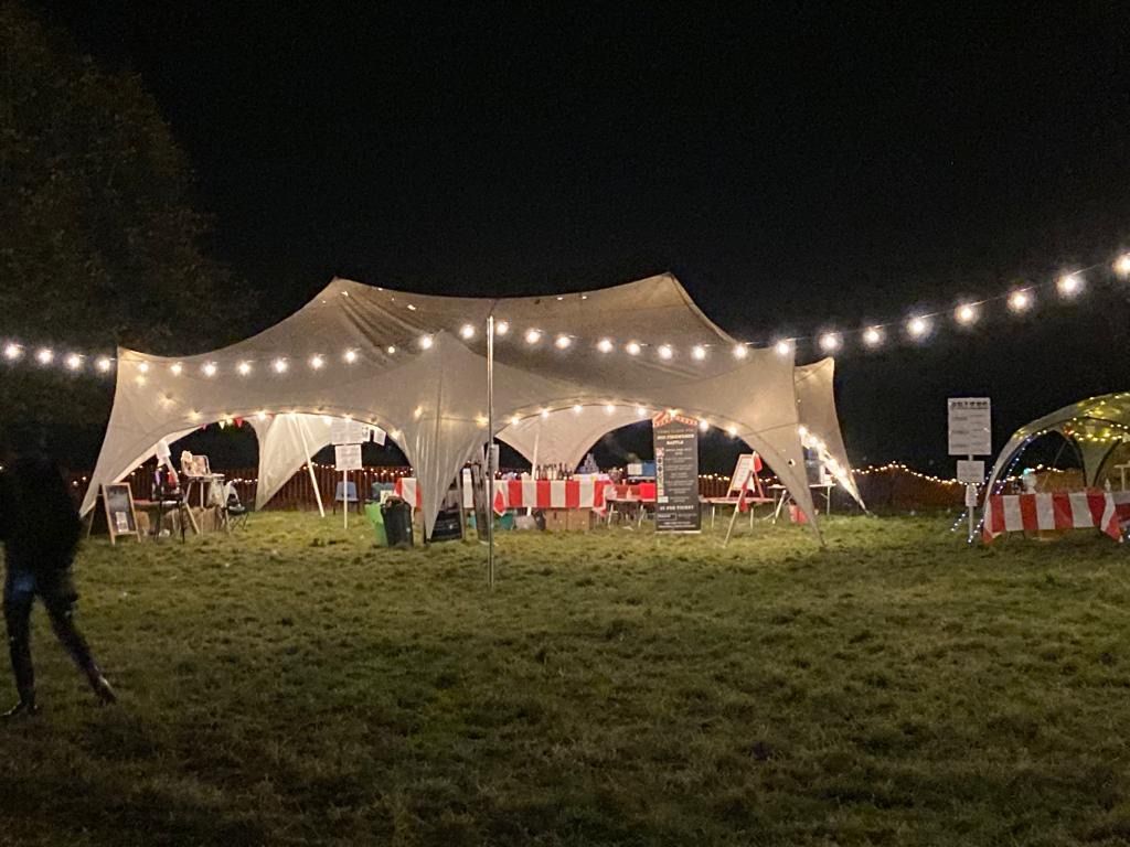 A nighttime outdoor event. Lit tent with string lights, tables, and a person in the foreground.