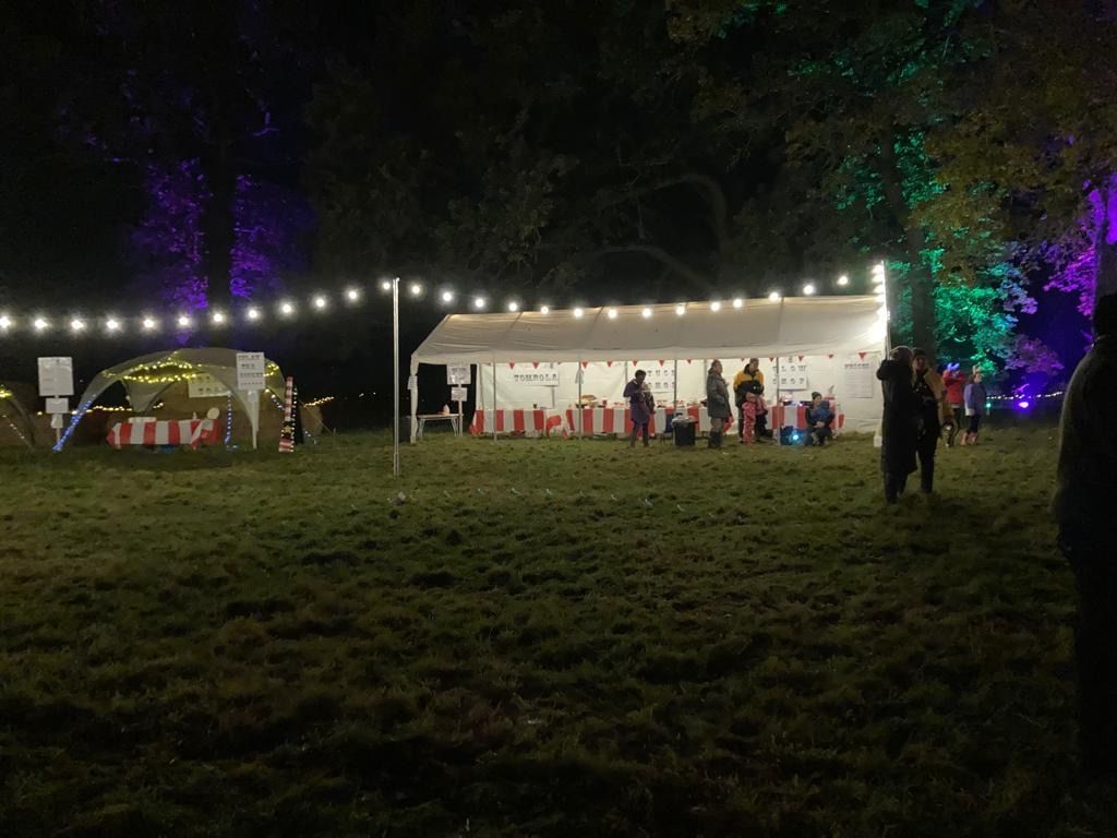 Night scene of a food tent lit with string lights in a park. People gathered, trees lit in purple and green.