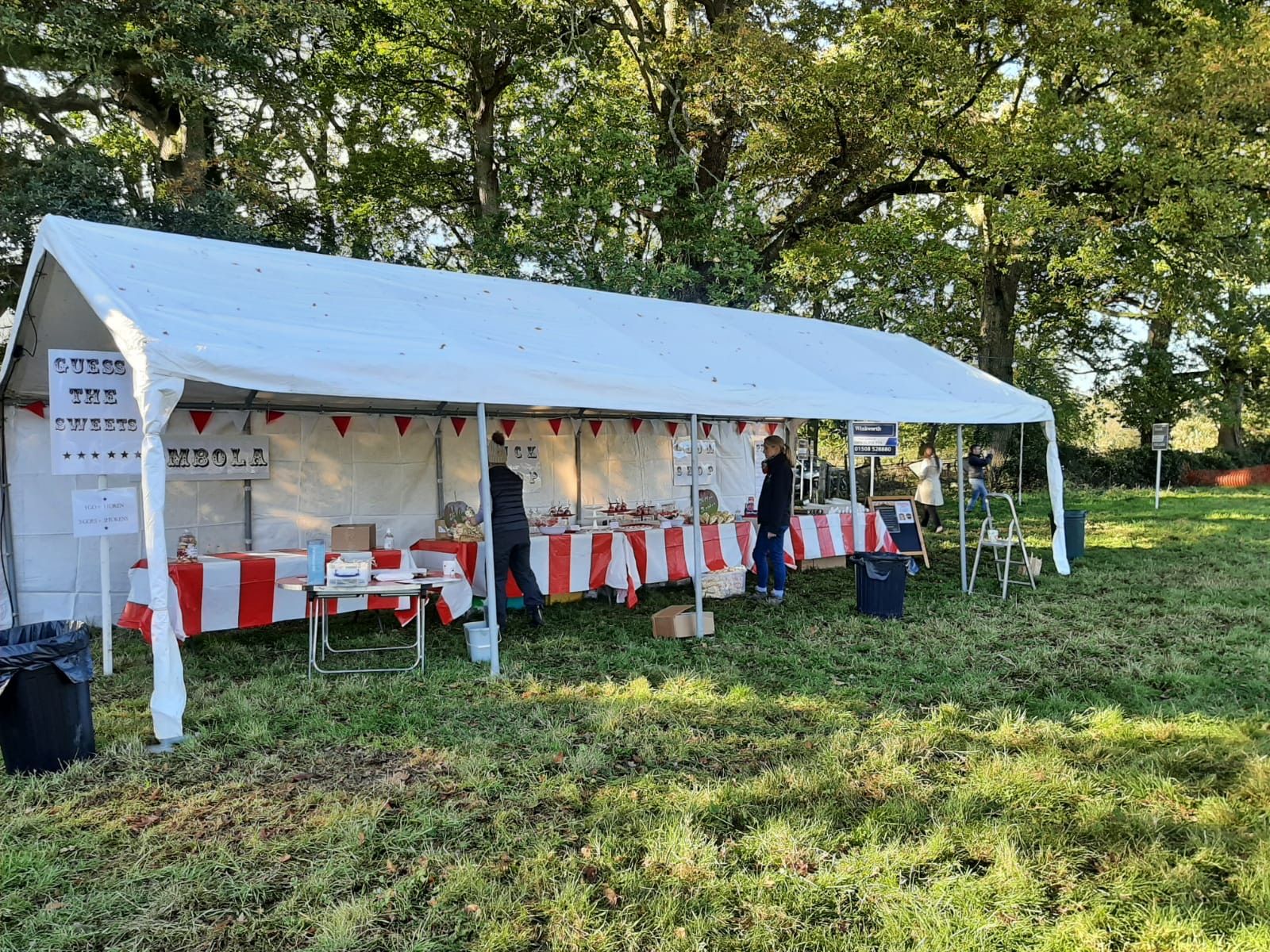 White tent with red and white tables set up outdoors, people at a food stand.