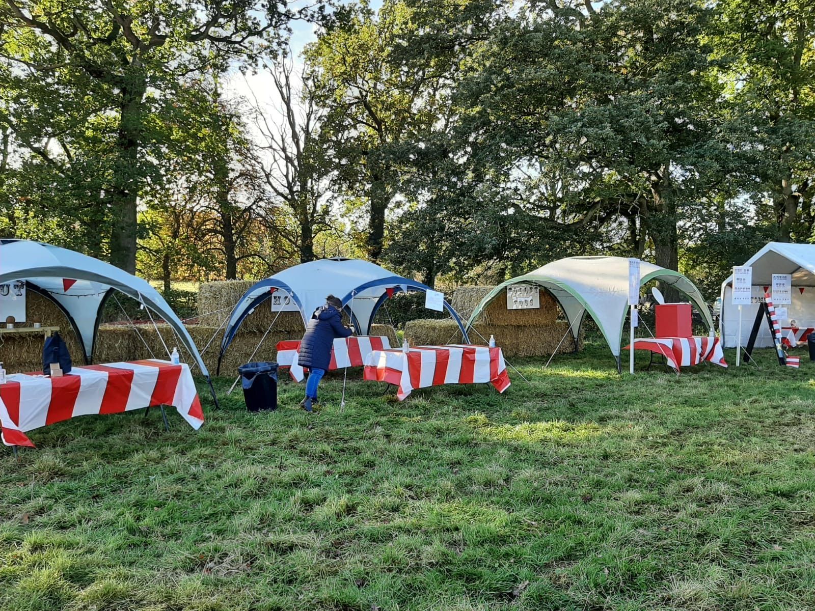 Outdoor market stalls with red/white tablecloths and canvas tops in a grassy area, trees in background.