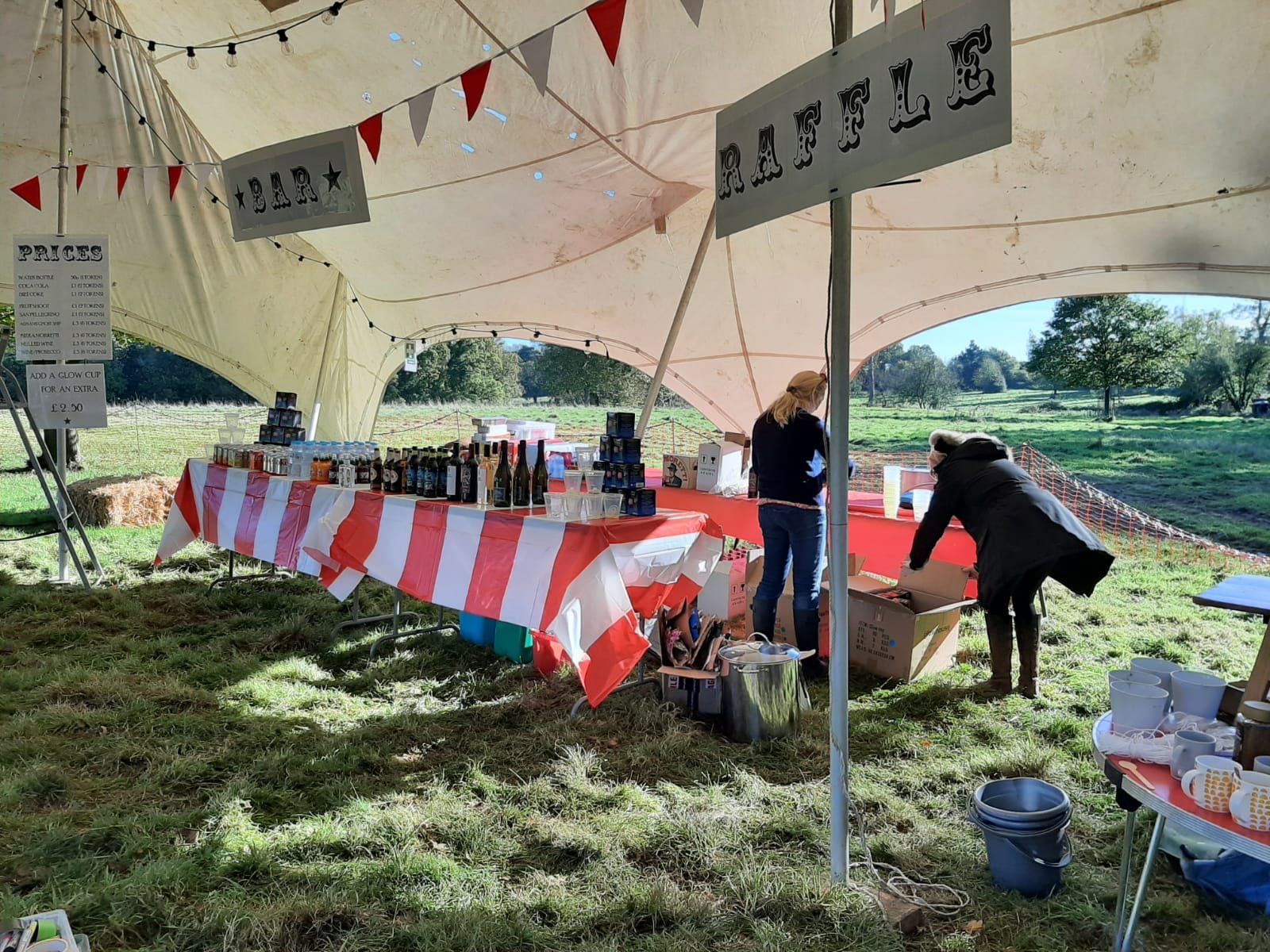 A white tent with a red-and-white striped table displaying goods. Two people are behind the table in a grassy field.