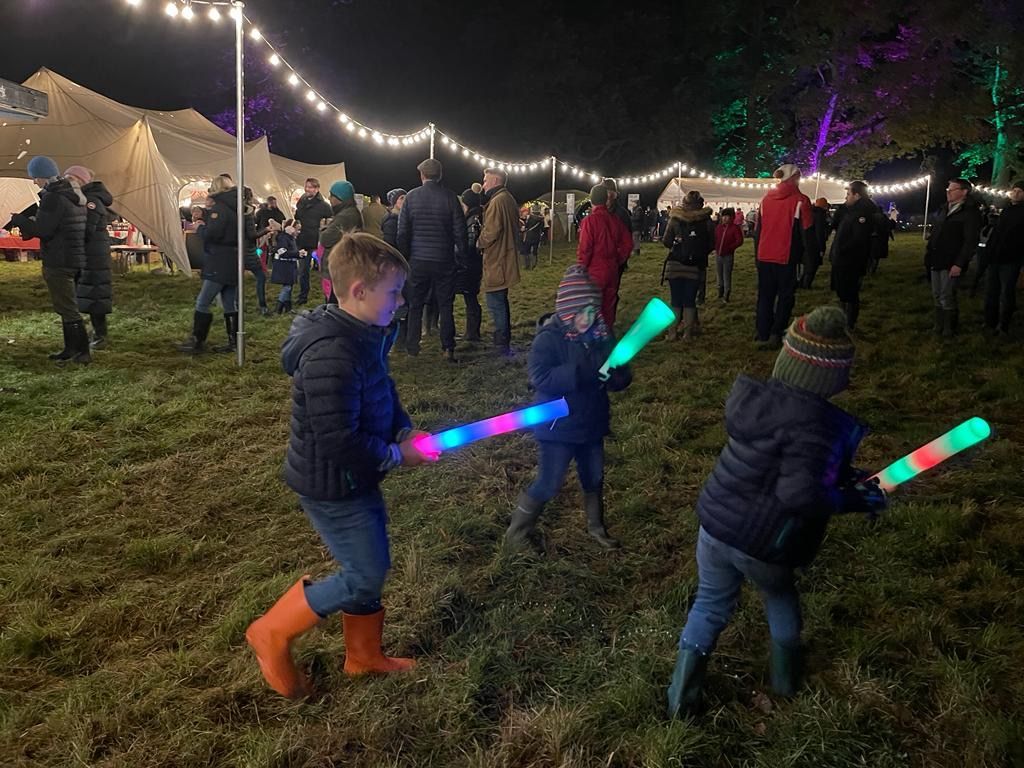 Children playing with light-up toy swords at an outdoor evening event with tents and string lights.