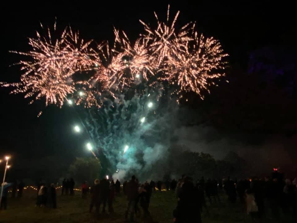 Fireworks burst in the night sky above a crowd of people gathered on a grassy field.