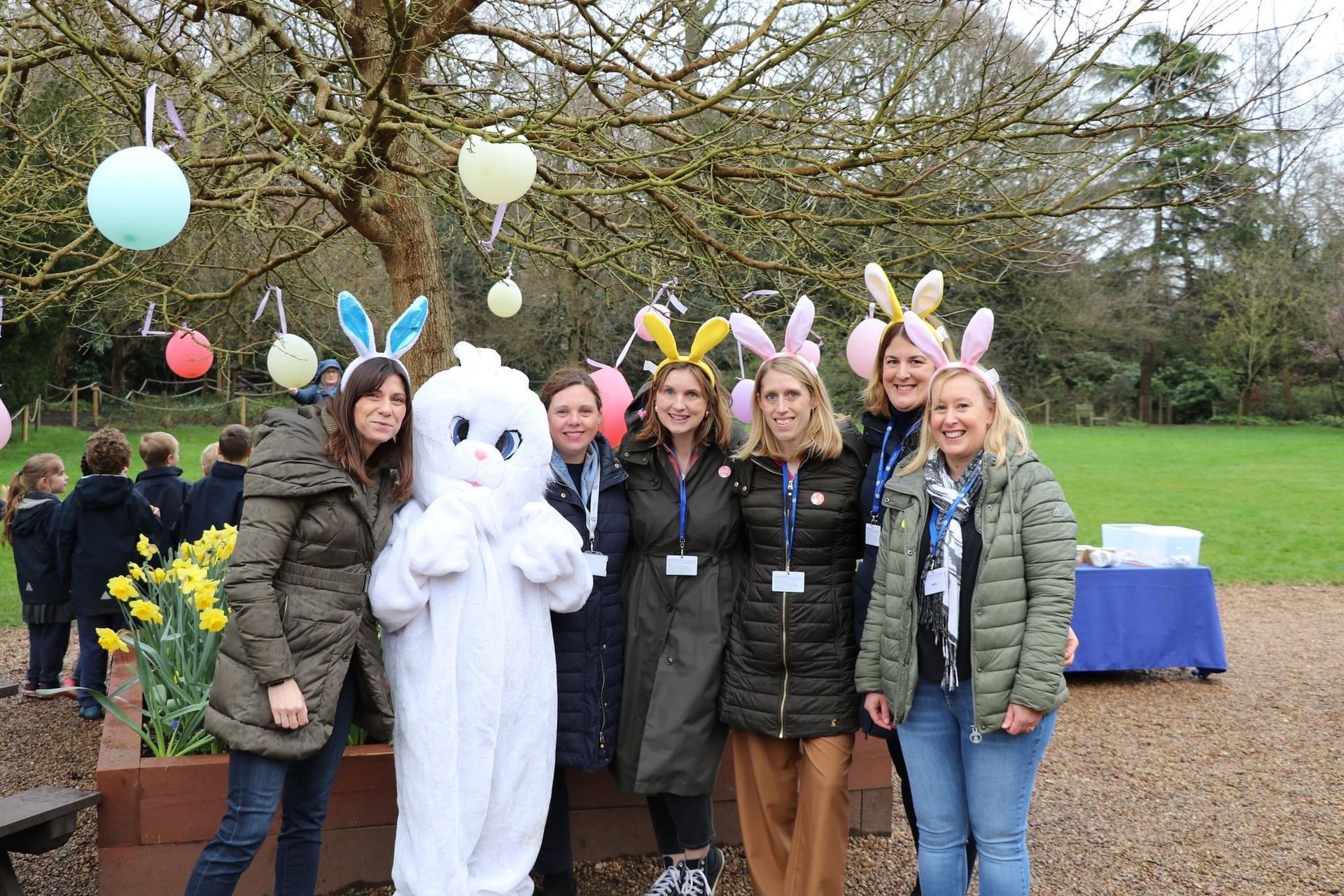 Group of people wearing bunny ears, posing with Easter Bunny outdoors, near tree decorated with hanging eggs.