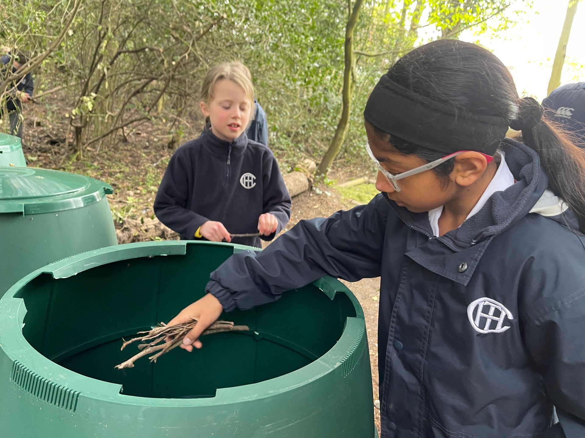 Two students in blue uniforms examine sticks inside a green bin outdoors.