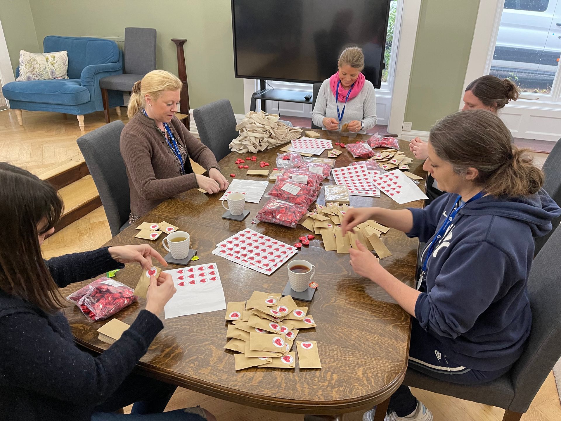 Five women seated around a table are packaging items; brown bags, red hearts, and cards are visible.