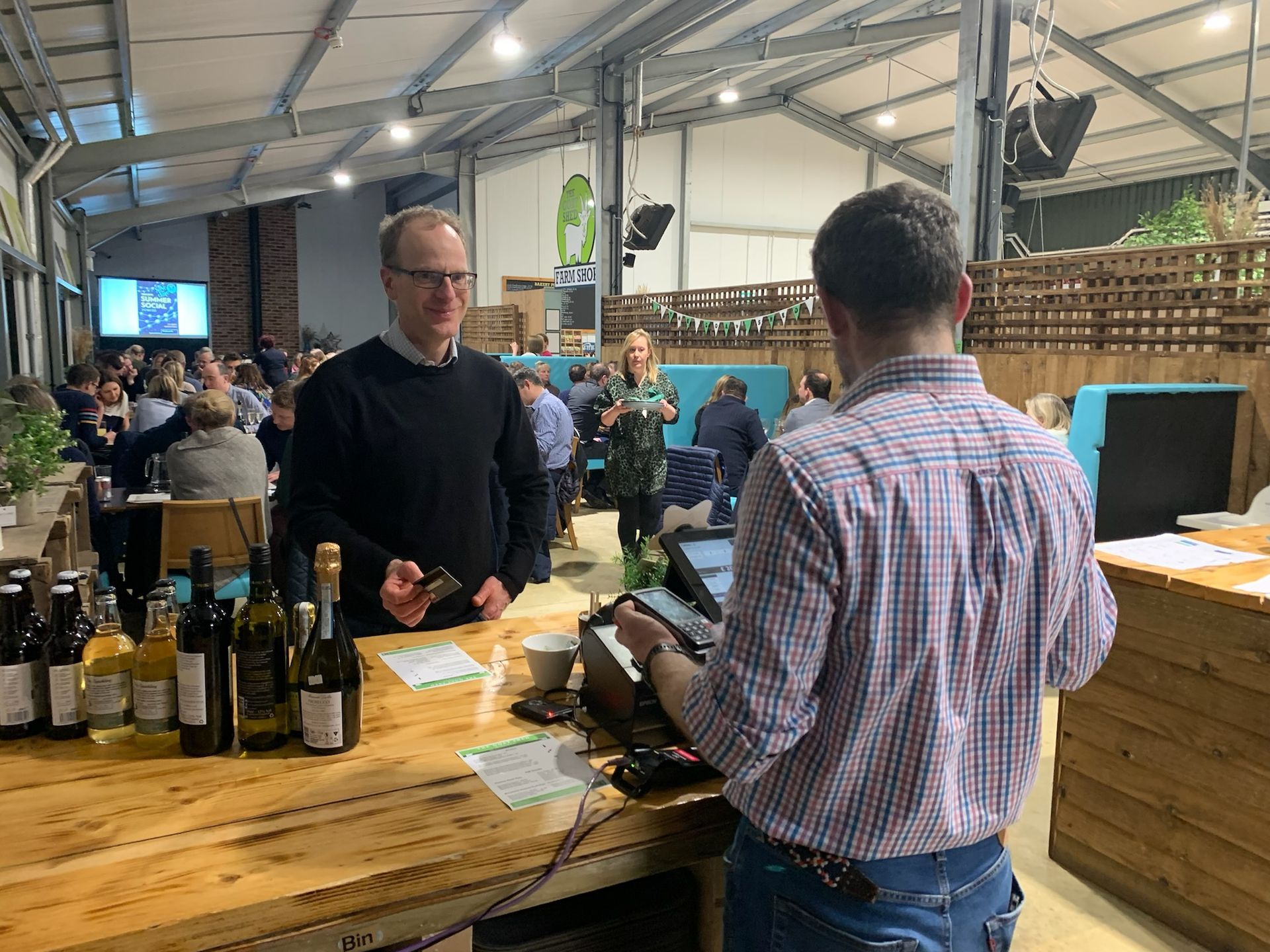 Man at bar being served at an event; various wine bottles, people in background.
