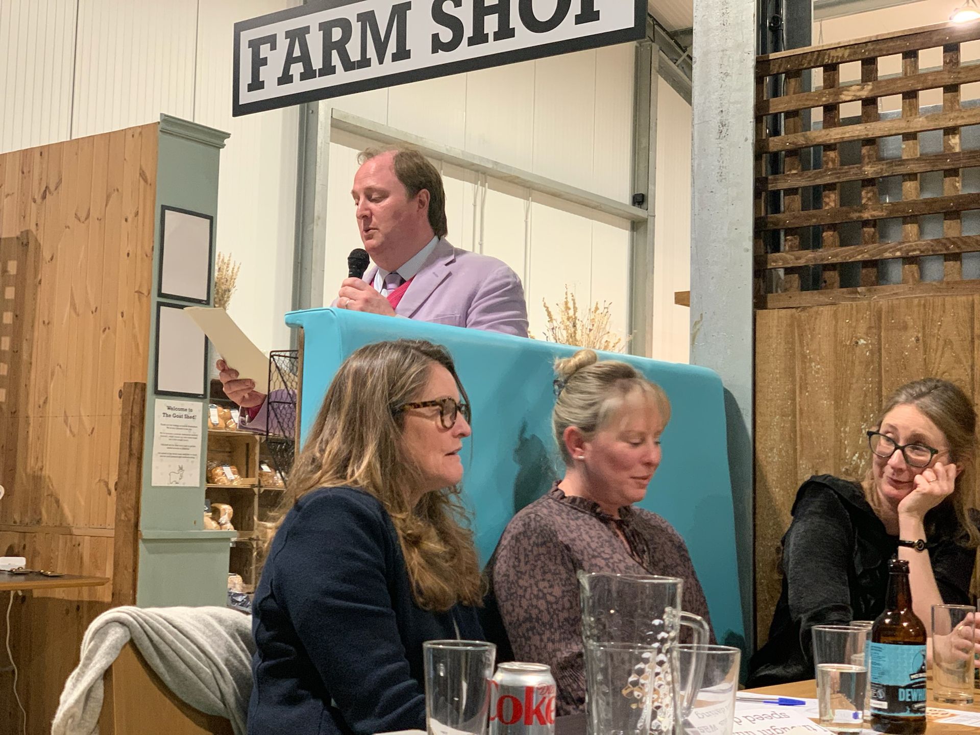 A man with a microphone speaks at a farm shop auction. Women seated at a table listen.