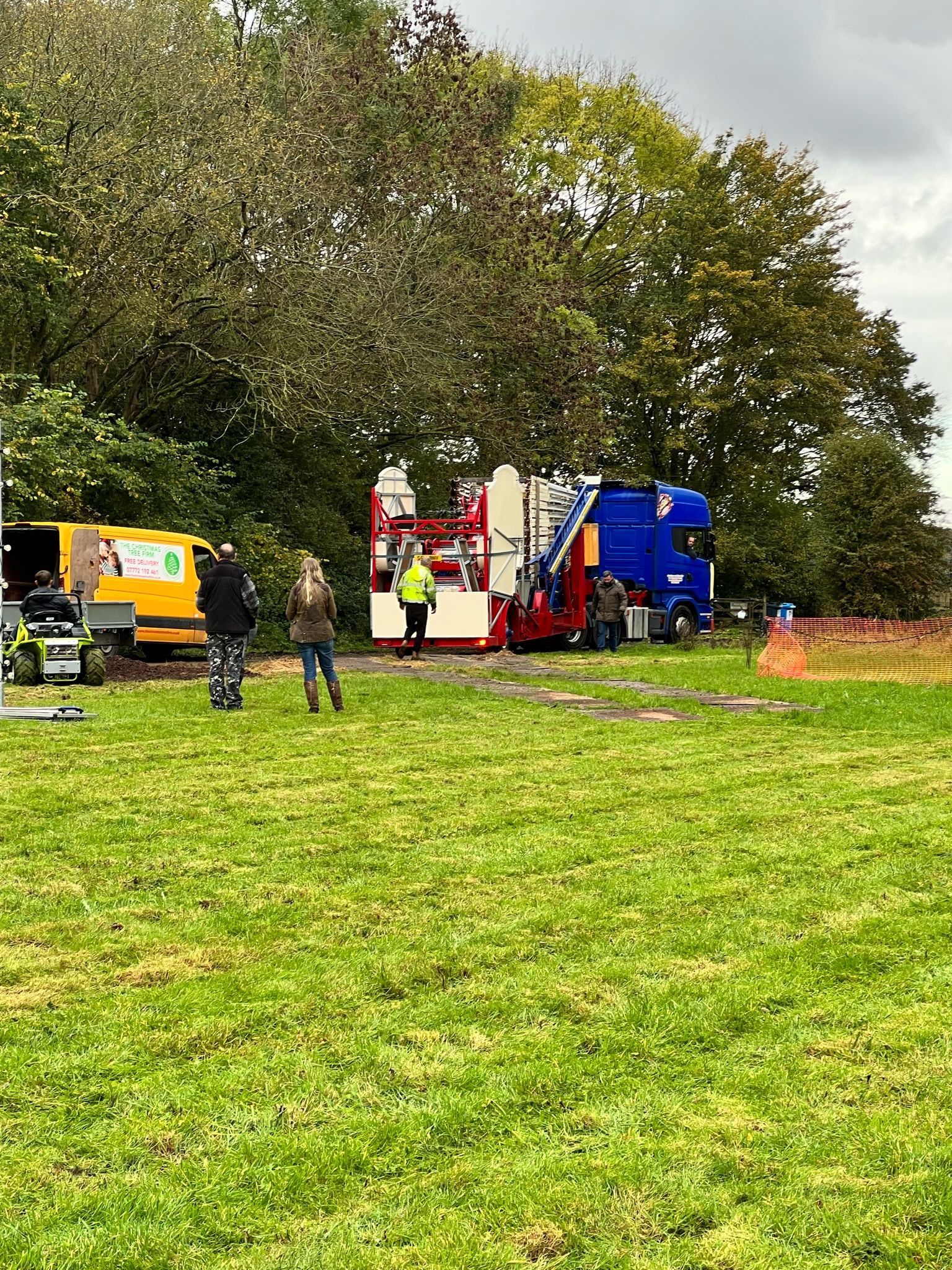 People near a red and blue truck, yellow van, in a grassy field with trees.